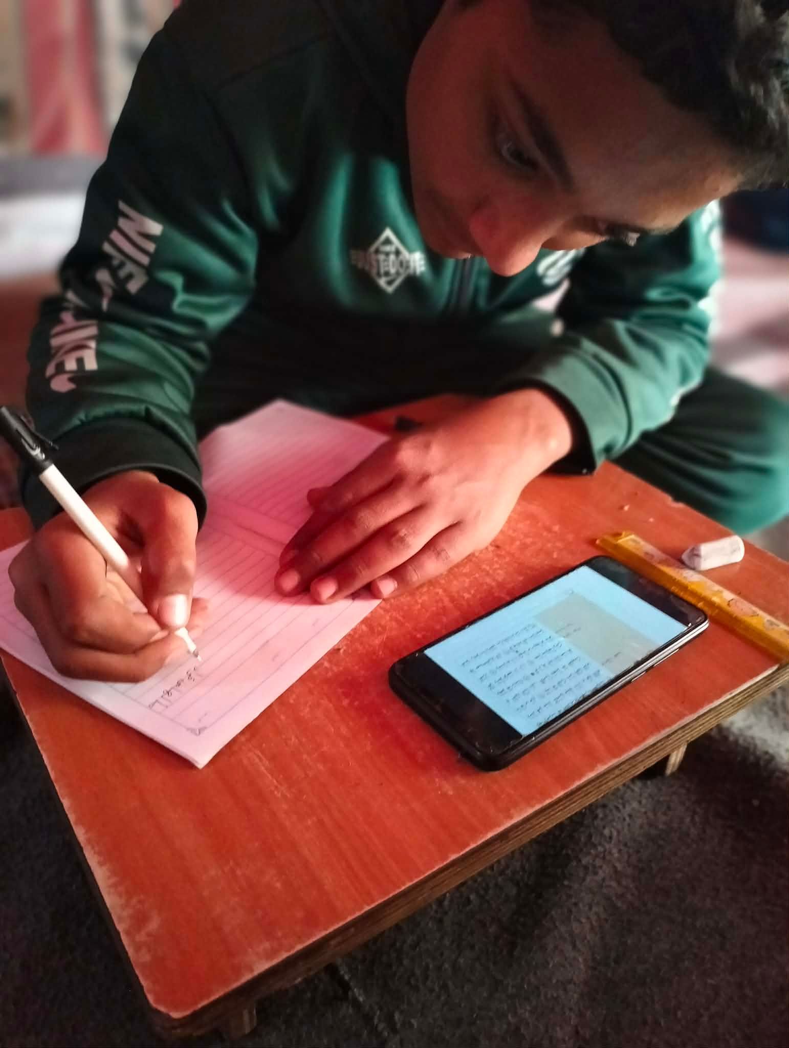Abdul Rahman in a green sweatshirt writing on lined paper with a pen, a smartphone with text on the screen, a ruler, and an eraser are on a wooden desk.