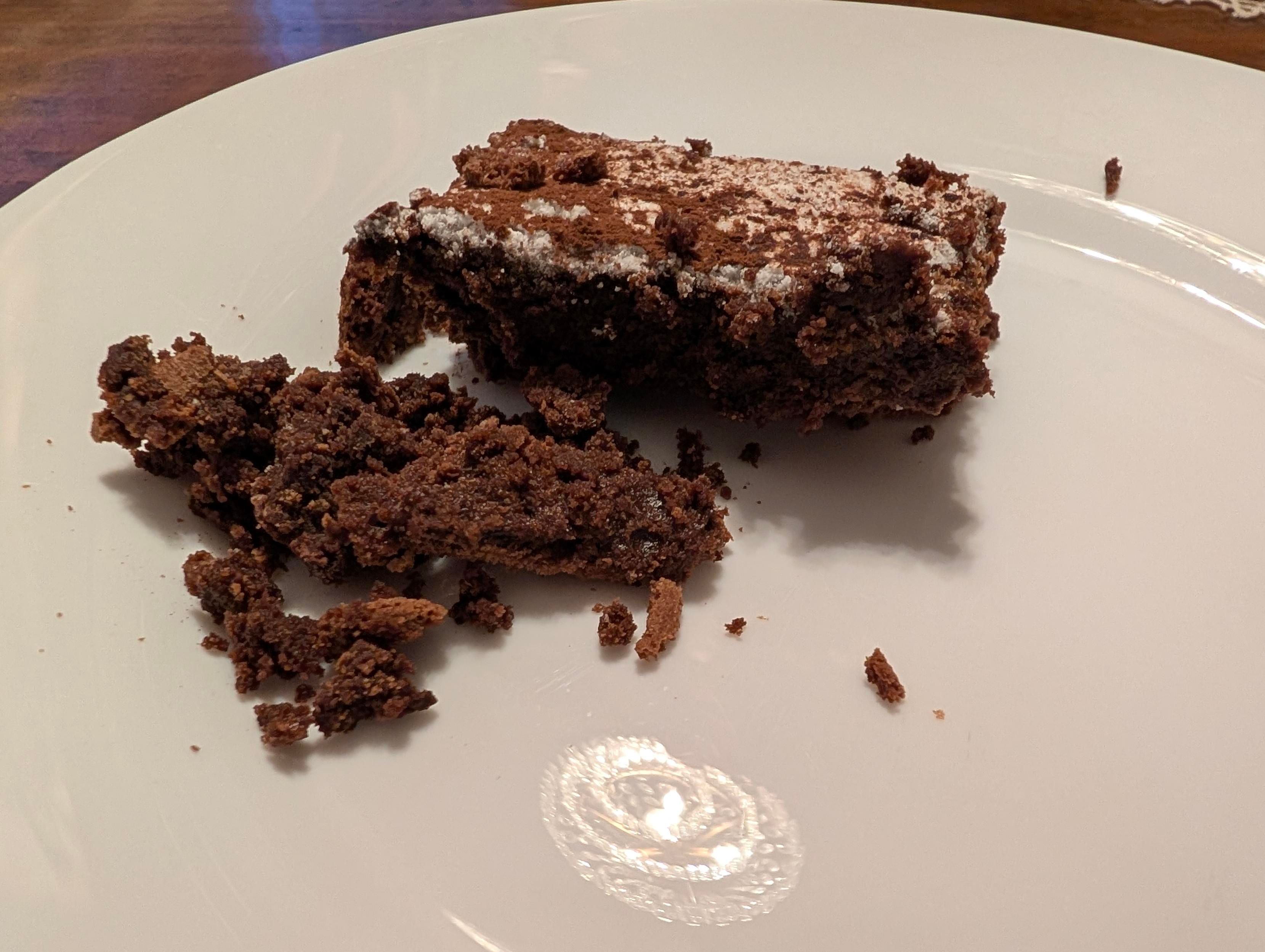 A partially eaten slice of tenerina, a dense chocolate cake, sits on a white plate with loose crumbs scattered beside it and a light dusting of powdered sugar on top.