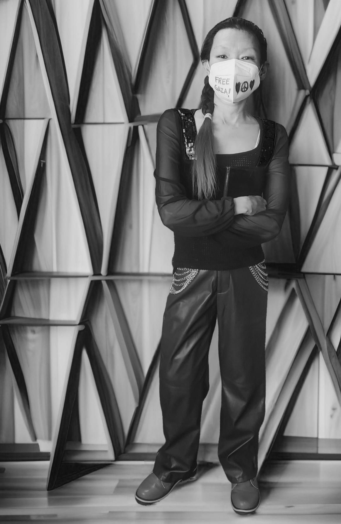 Black and white full-length portrait of a woman(me) in front of an artistic geometric wall. She wears a face mask with “FREE GAZA!” written on it, black top with sequin shoulder detail, black pants with chain trim, and dark shoes. Her arms are crossed, and she has her hair in a side braid.
