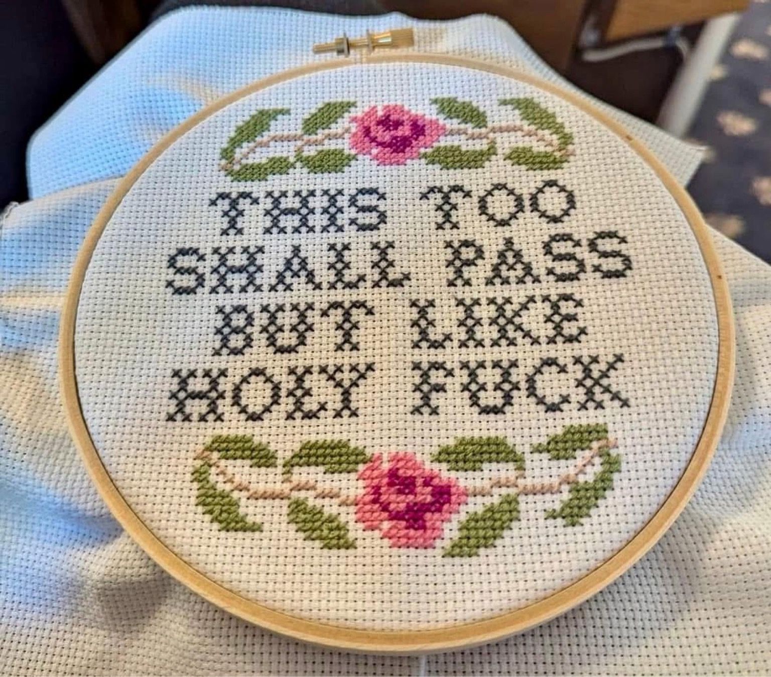 An embroidery hoop showcases flowers with the text: THIS TOO SHALL PASS BUT LIKE HOLY FUCK. 