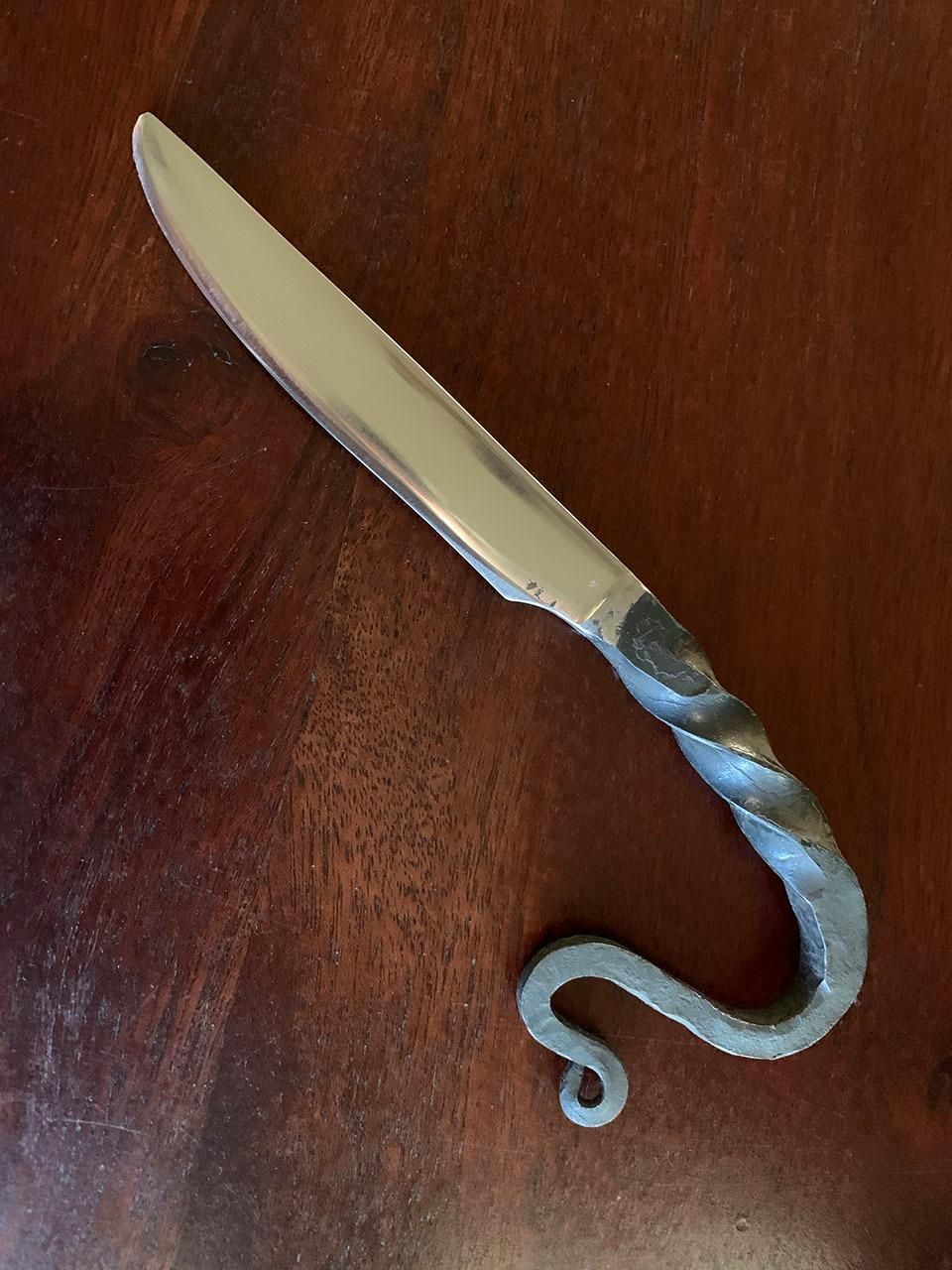 A blacksmithed butter knife with a snake- or dragon-like tail. The blade section is polished but the handle section has been left with a dark "blacksmith finish."