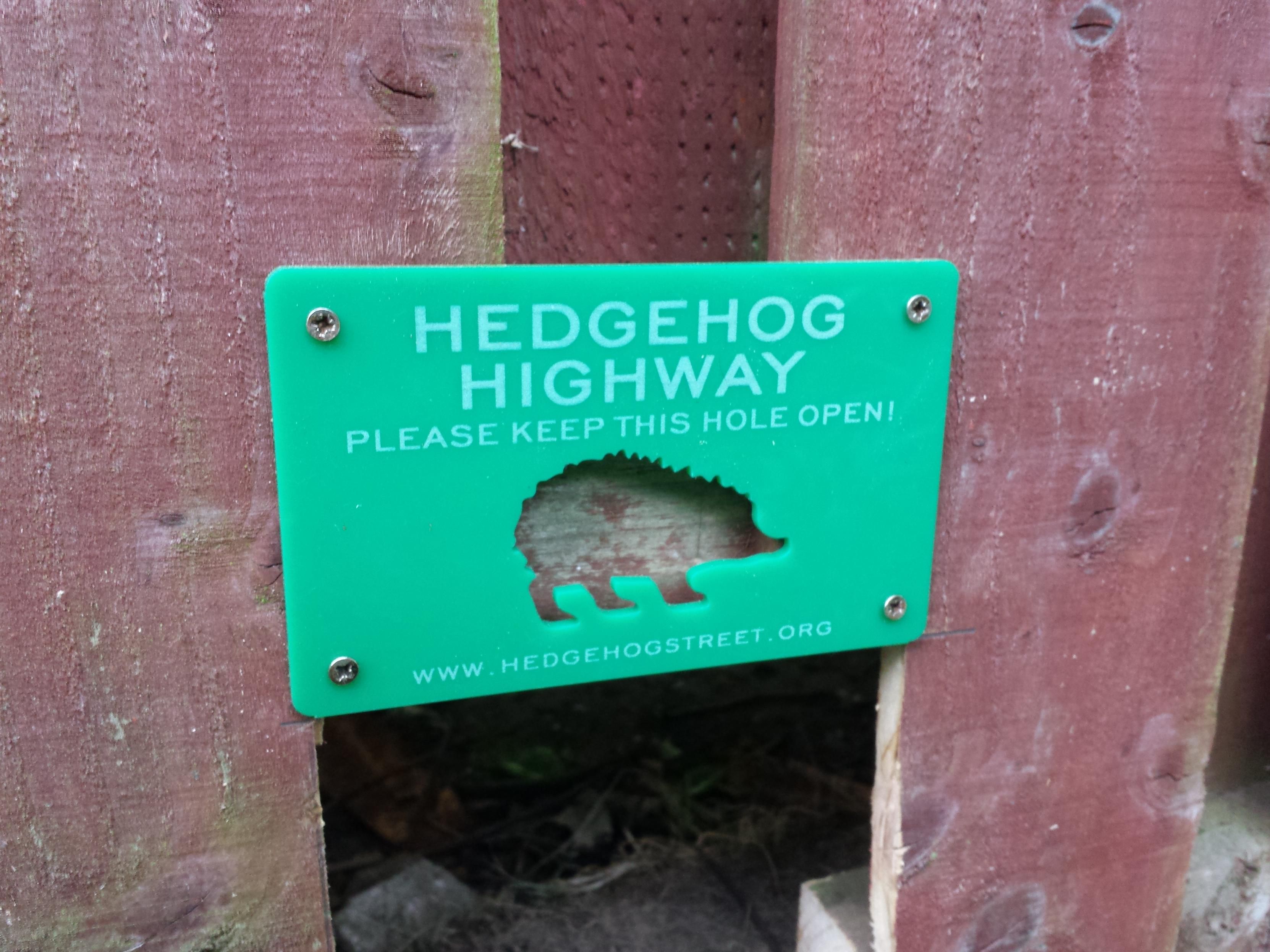 Close-up photo of the green plaque. It has a cutout in the shape of a hedgehog and reads: Hedgehog highway. Please keep this hole open. Www.hedgehog street.org