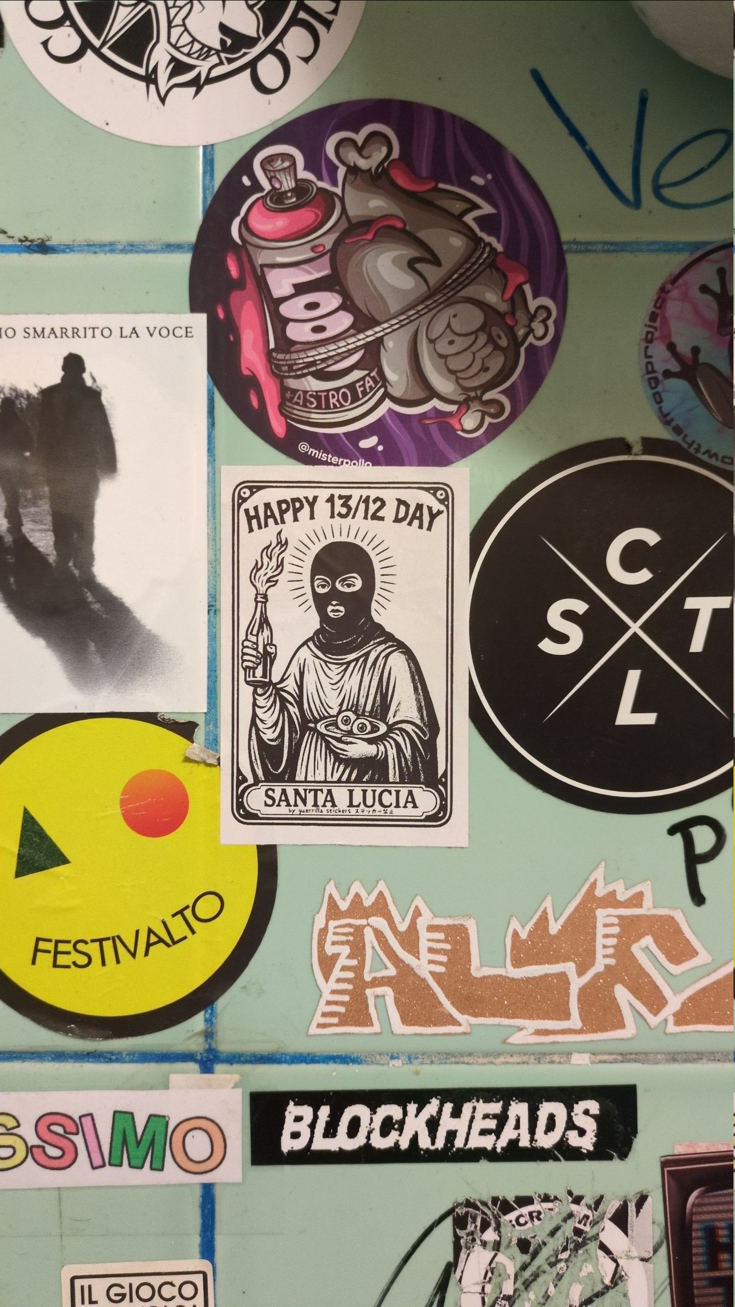 Parete del bagno di un locale rivestita di piastrelle verde chiaro e coperta da numerosi adesivi. Al centro uno sticker in bianco e nero con la scritta “HAPPY 13/12 DAY – SANTA LUCIA”, raffigurante una figura incappucciata con aureola che tiene una bottiglia incendiaria e un piattino con 2 occhi. Attorno altri adesivi e illustrazioni colorate.