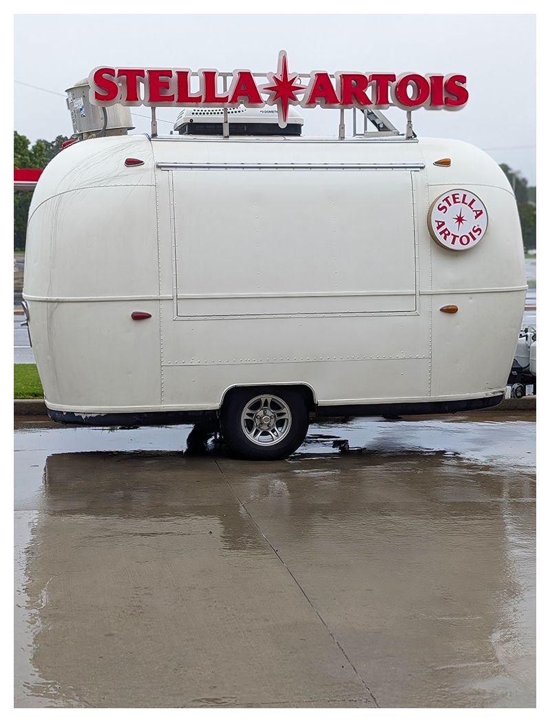 ai-assisted alt text:

A small, white vintage trailer with a Stella Artois logo on the side is parked on wet pavement. Above the trailer is a large, red “STELLA ARTOIS” sign with a star. The ground appears wet, reflecting part of the trailer and sign.