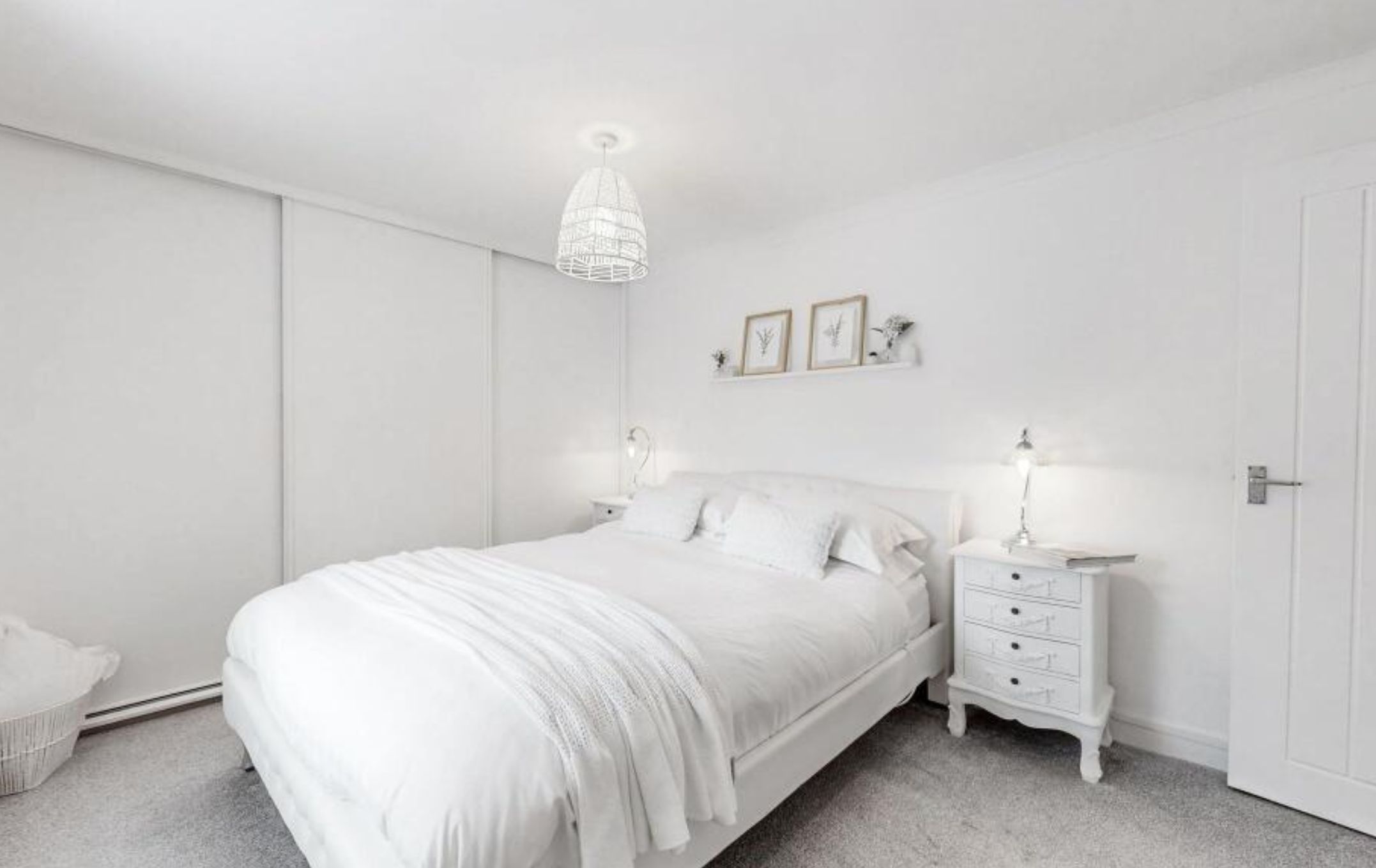 A bedroom with light grey carpet and bright white walls, ceiling, closet, bed, bed linens, ceiling lamp and side table. There are 2 small pictures on a shelf above the bed, each has a leaf. 