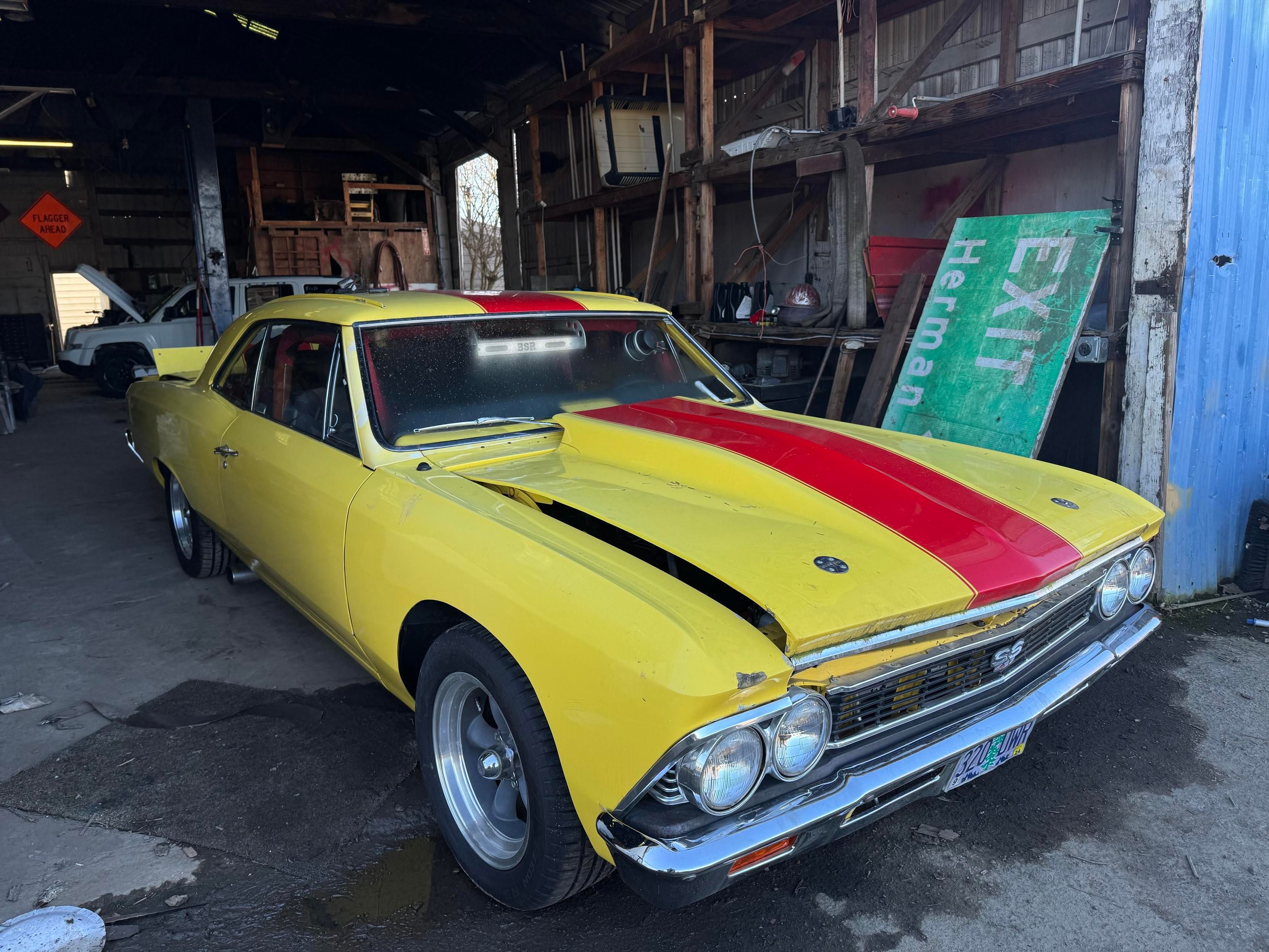 A yellow muscle car with a red stripe is parked in a barn-like setting. The car shows signs of aging and damage, with a partially open hood and a dusty exterior. Behind it, there's a green "EXIT" sign and various tools and equipment