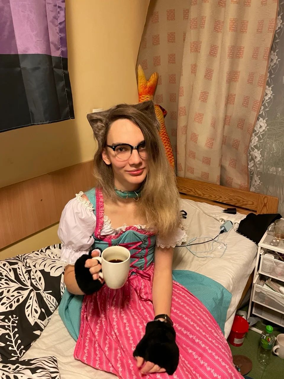 Me sitting on a bed with a coffee mug in a white, pink & light blue dirndl, cat ears, paw gloves and a black pair of glasses. 