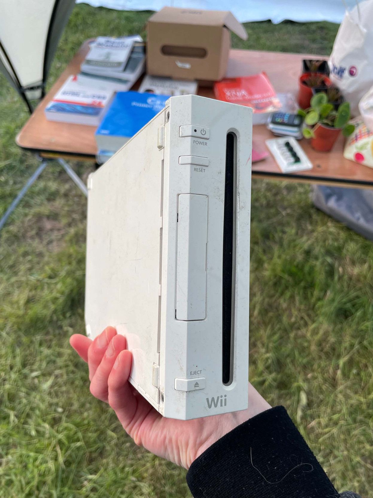 Picture of someone holding up a fairly scruffy Nintendo Wii. It's at the EMF 2024 Swap Shop, so the image is taken in a field with a table behind with an assortment of various Swap Shop goodies on it, including tech books, an Oculus dev kit, and what appears to be a venus flytrap.