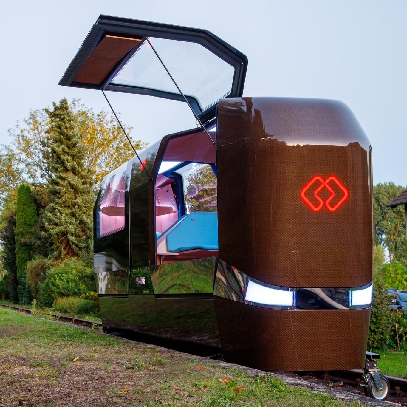 This image shows a gyro-stabilized monorail cabin. The cabin has a reflective, metallic exterior with large windows on the sides with one of them being open and revealing a cozy interior with seating and wood accents. It is situated outdoors on a patch of grass, with residential houses in the background and a clear sky overhead. 