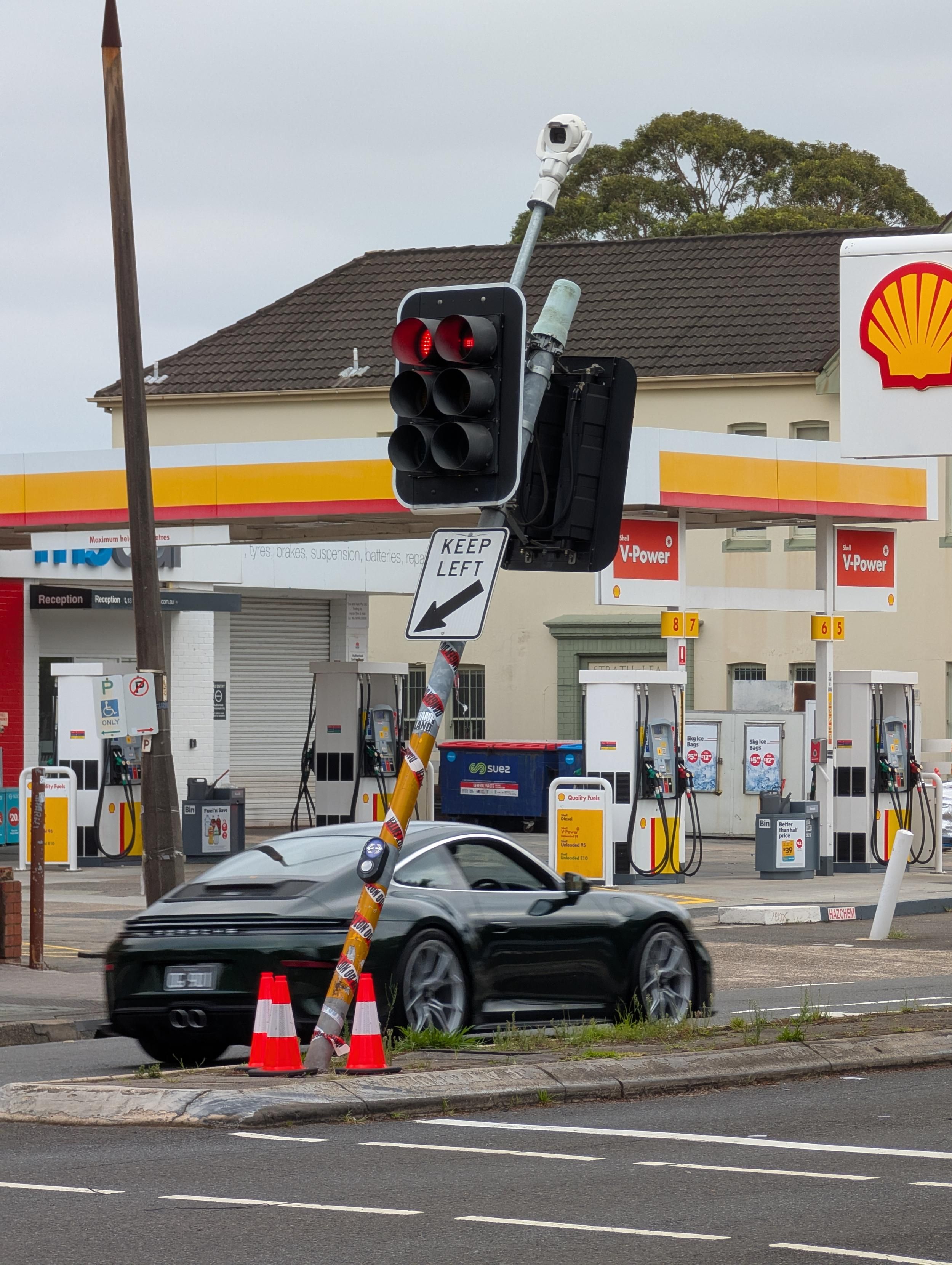 This image shows a street intersection scene with a Shell gas station in the background. A traffic light displaying red lights is mounted on a pole that also holds a sign reading "KEEP LEFT" with a left-pointing arrow; the pole is wrapped in stickers. In the foreground, two orange traffic cones are positioned near the base of the pole, and a black sports car is on the road. The gas station features yellow and red branding, multiple fuel pumps with "V-Power" signage, and a building with a "Reception" sign and closed shutters. A blue container and a tall pole are also visible in the scene.