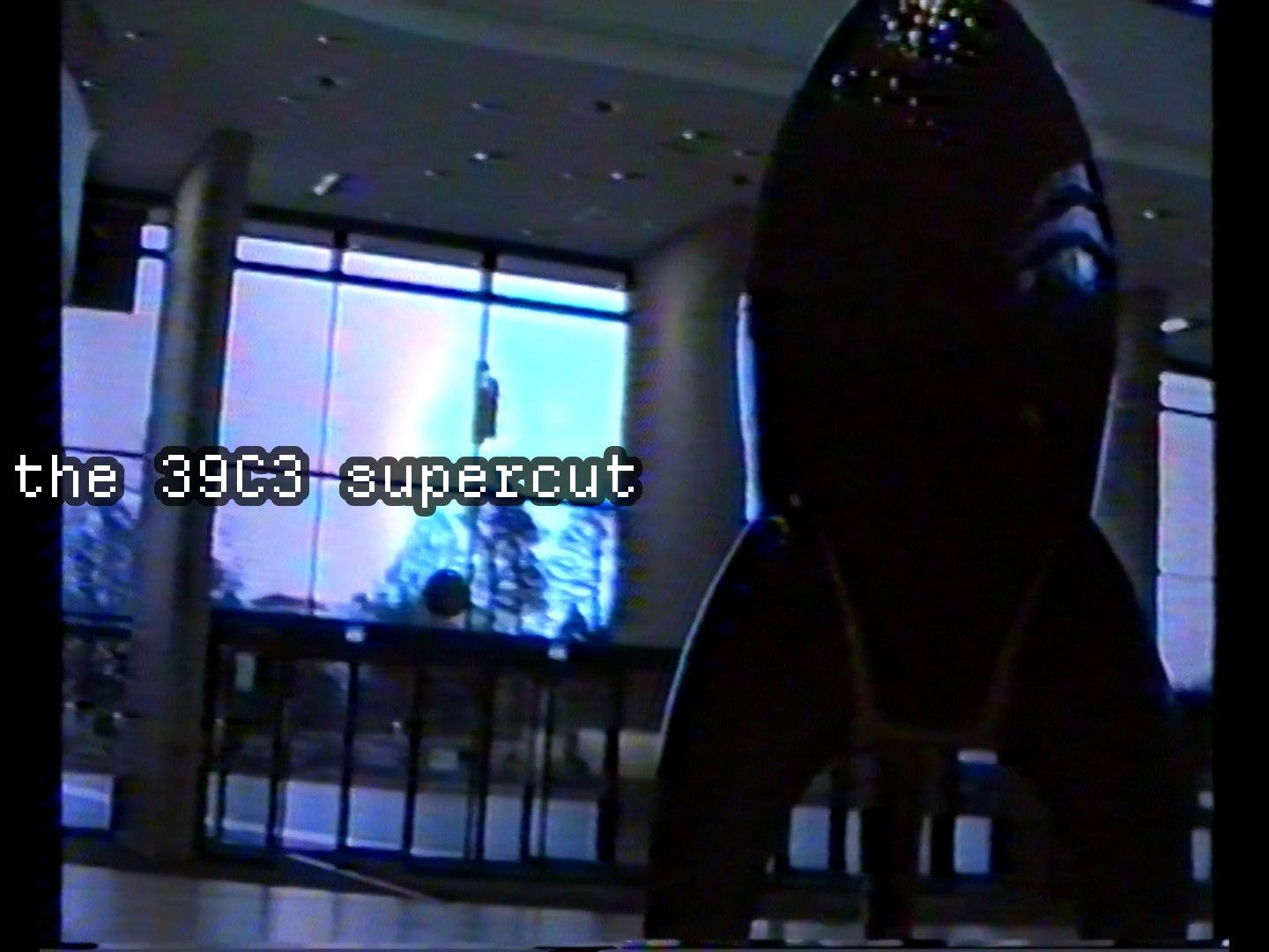 a dark frame from the video; on the left, monospace pixel font says "the 39C3 supercut". the background is the bottom floor of the main entrance to CCH during congress; on the right, the large fairydust rocket dominates the frame.