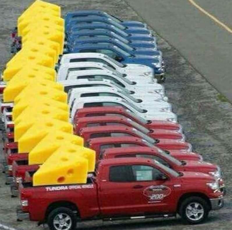 Photo of 18 pickup trucks in grouped colors of red, white, and blue all parked in a row. In the back of each is a large wedge of cheese.