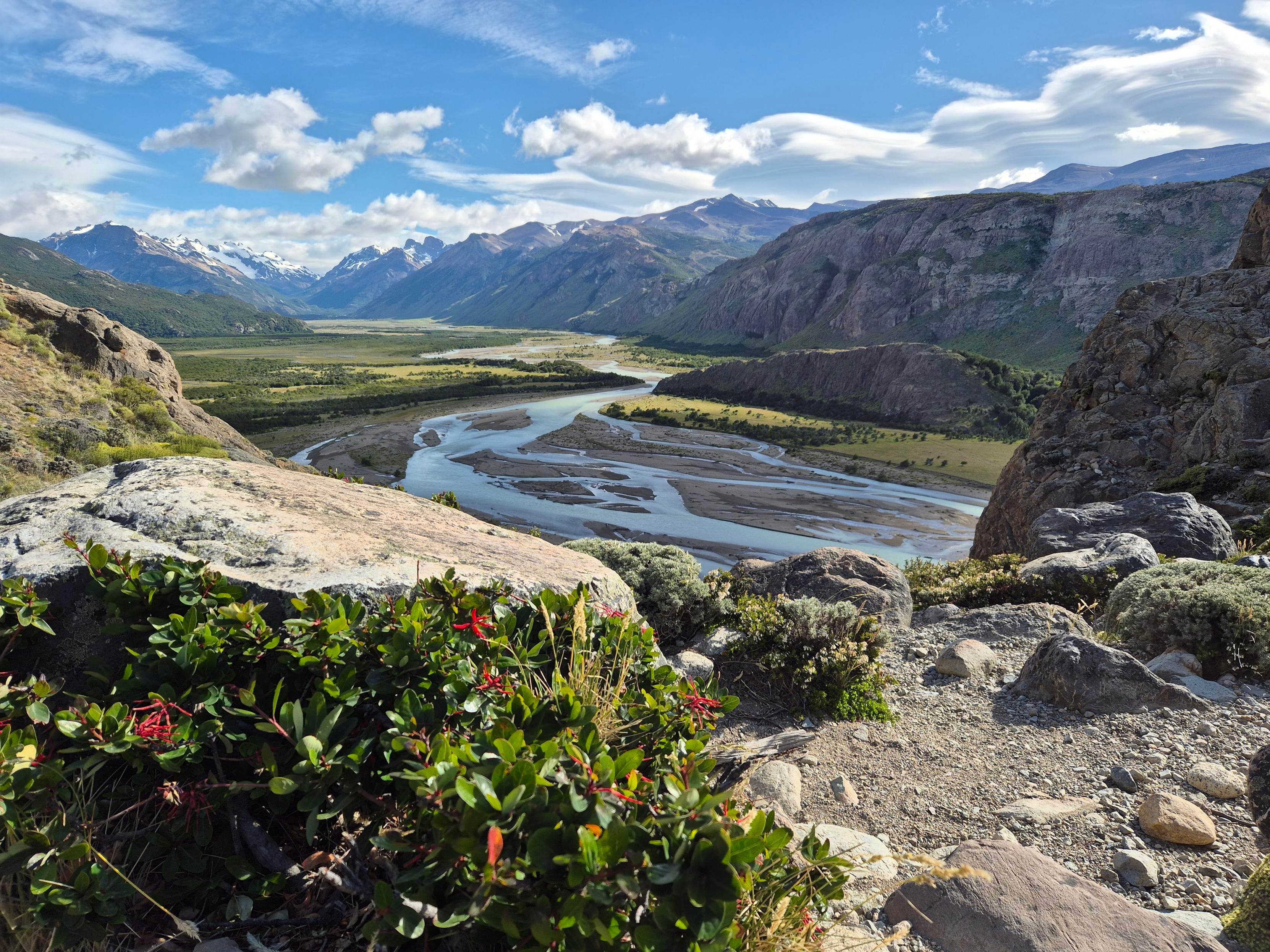 View down into a beautiful broad valley surrounded by stip high mountains with a broad river wirh multiple arma flowing through it. 