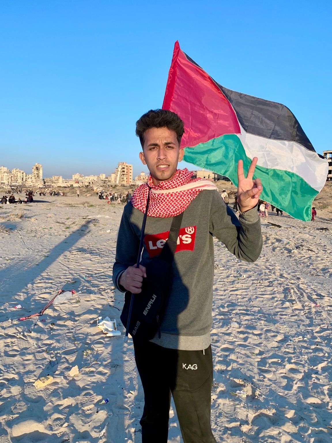 Mohammed, 22, from Gaza, standing before the ruins of Gaza City, holding a Palestinian flag, doing a peace sign with his fingers, looking tired and thinned out but positive and hopeful.