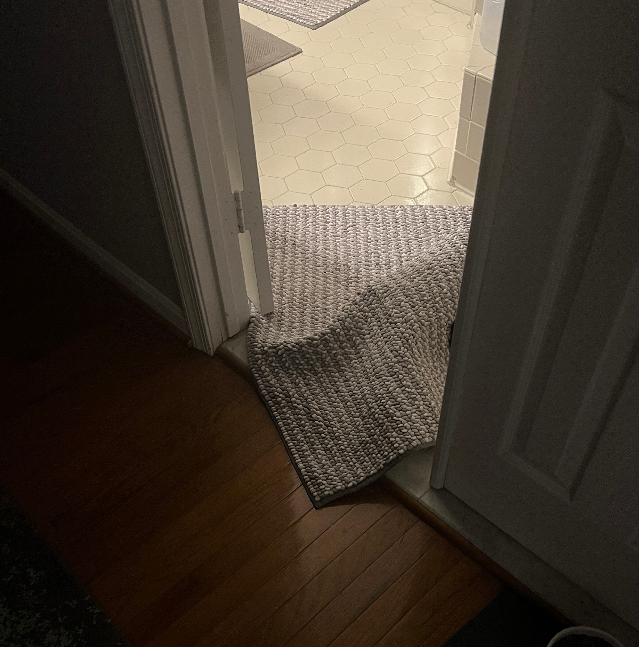 Photo of a grey knit bathmat quite lumpy and kinda blocking the doorway to the bathroom. 