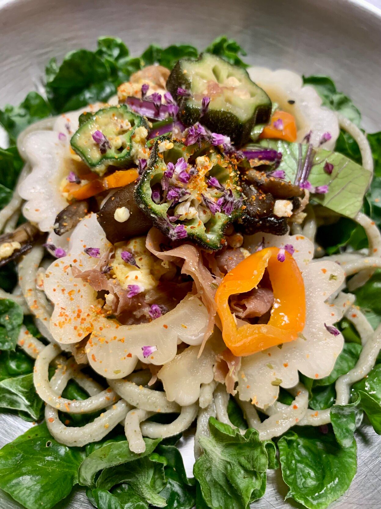 In a silver bowl on top of a bed of green curly sissoo leaves are light green thin jackfruit leaf soba noodles. On top of the noodles are slices of white bitter melon, maitake mushrooms, sliced mizuna leaves and dark pink stalks, fried egg, sliced stars of okra, prosciutto, orange slices of madhatter pepper and topping it all off, a sprinkling of tiny purple shiso flowers. 