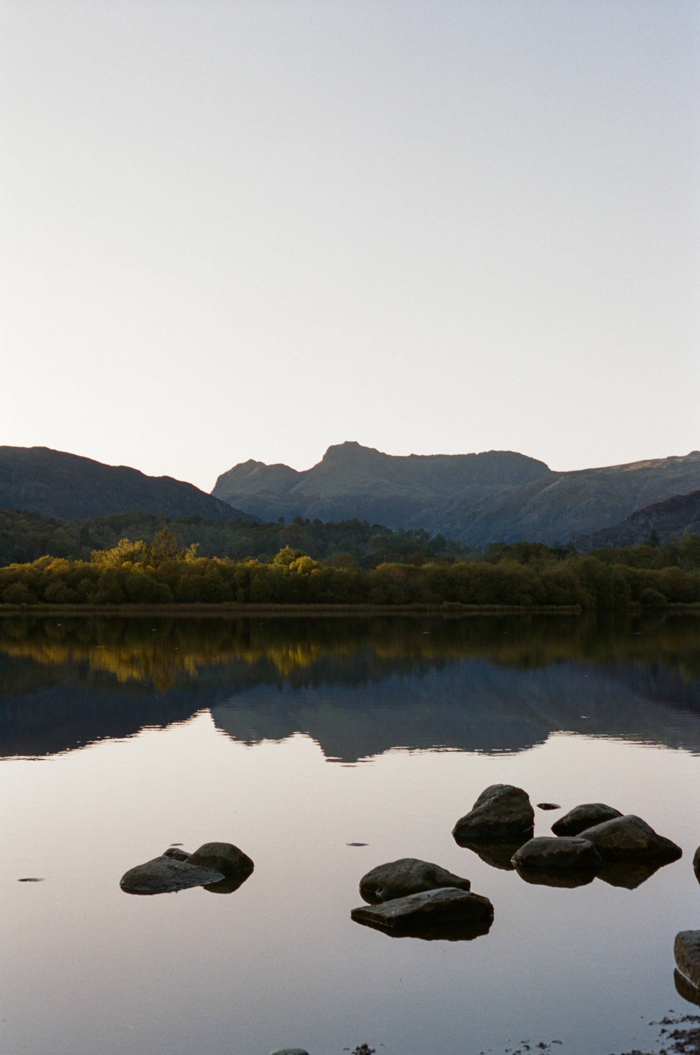 A calm lake, reflecting the auburn trees on the far shore and the Langdale pikes in the distance. Some rocks are in the water.