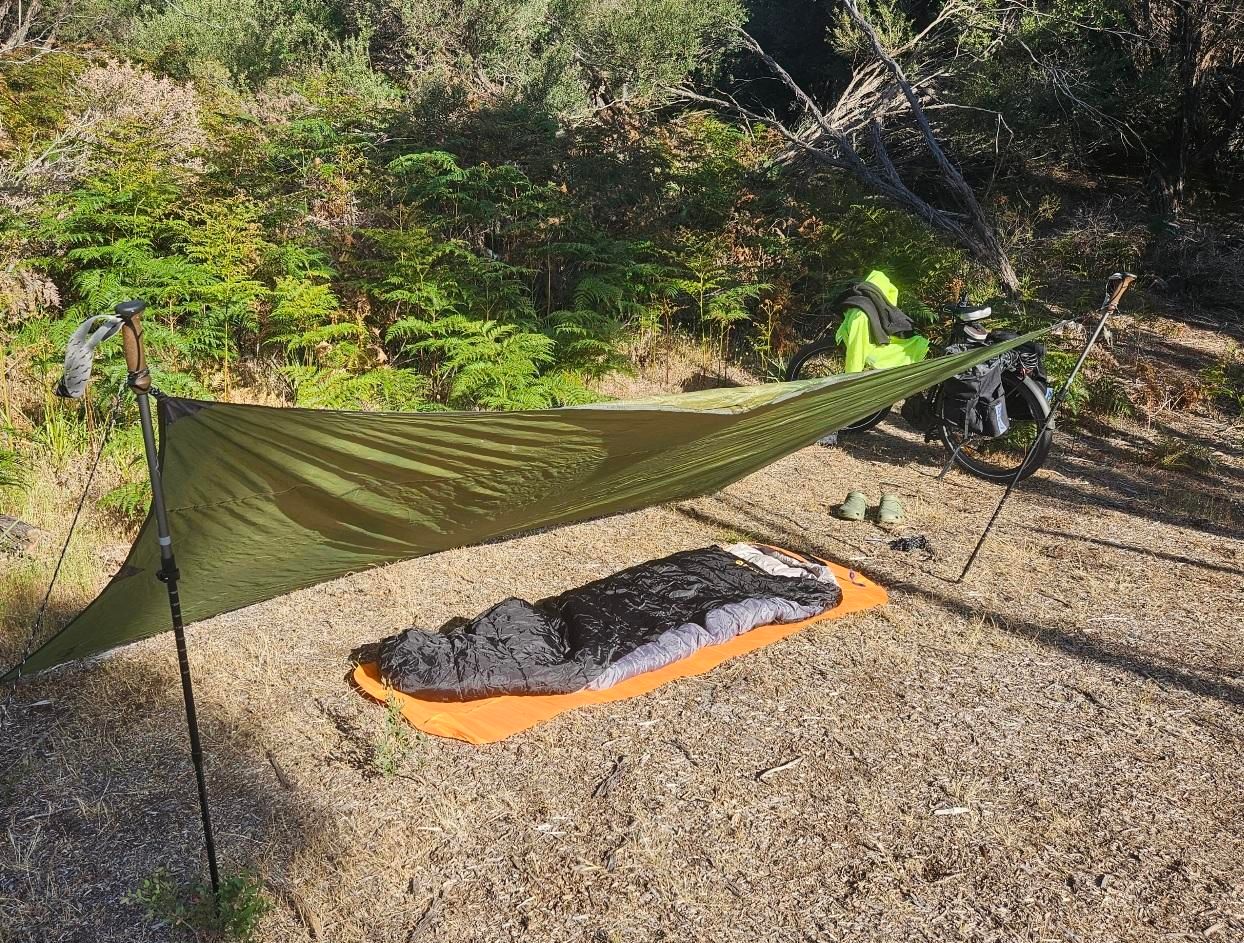My camp for two days: a tarp stretched between two hiking poles, and an inflatable mattress and sleeping bag underneath. In the background is my ebike, still to have its cooking gear unpacked, and a whole load of bracken and tea tree.