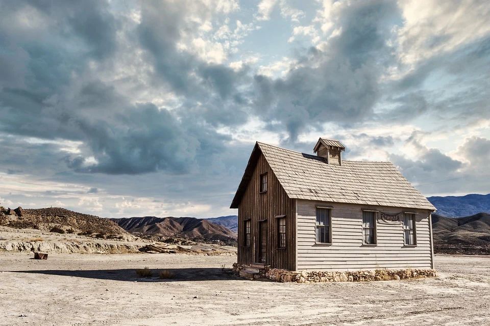 A rickety wooden house sits on a flat dirt plain, with some rolling hills and mountains in the distance. The sky is partly overcast and the tone of the image is cool icy blues and pastel browns.