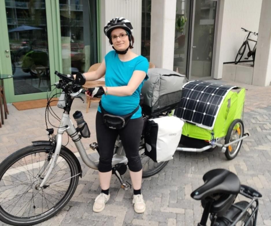 Me and my bike and trailer, standing in front of a building, on my way to WHY.
The bike, bags, and trailer look mostly the same as in the first picture, except the blue shopping bag is not there, and there's nothing attached to the outside of the trailer except for the solar panel (which also is attached in a much simpler way than before).
I am standing next to my bike, looking into the camera, wearing a cyan t-shirt, black leggings, a black fanny pack, beige shoes, fingerless gloves and a cycling helmet.