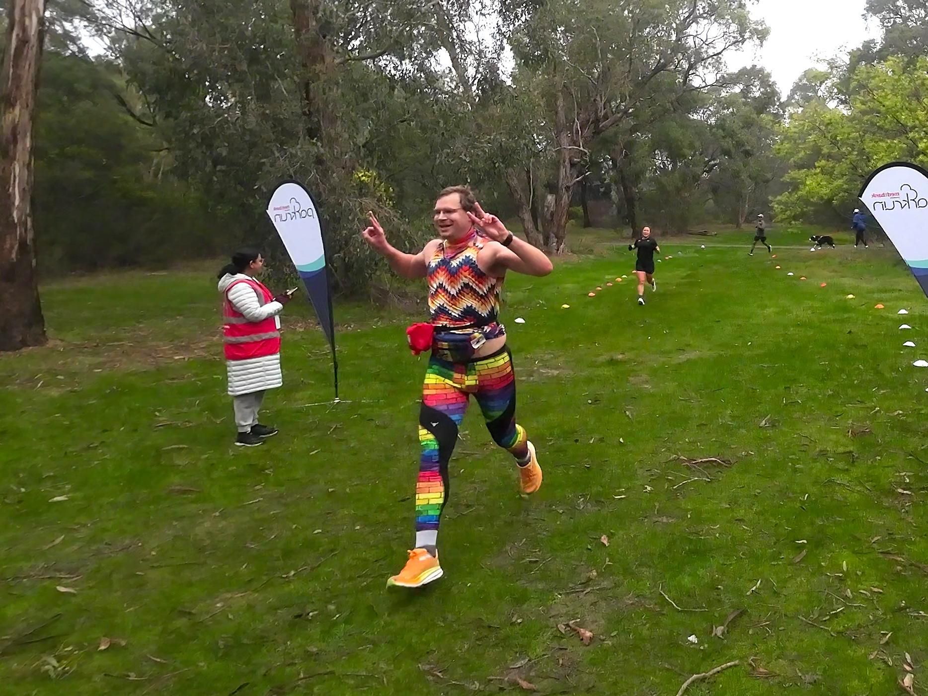 Picture of Luke as they cross the finish line at Parkrun. They are wearing their usual Parkrun getup - pride pattern leggings, colorful top, pride colored bandanna around their neck, pride patterned hip bag plus first aid kits around their waist.

The picture was snapped mid stride, with their front foot just about to come down as the back leg is lifting.

They are throwing Vs and smiling widely.