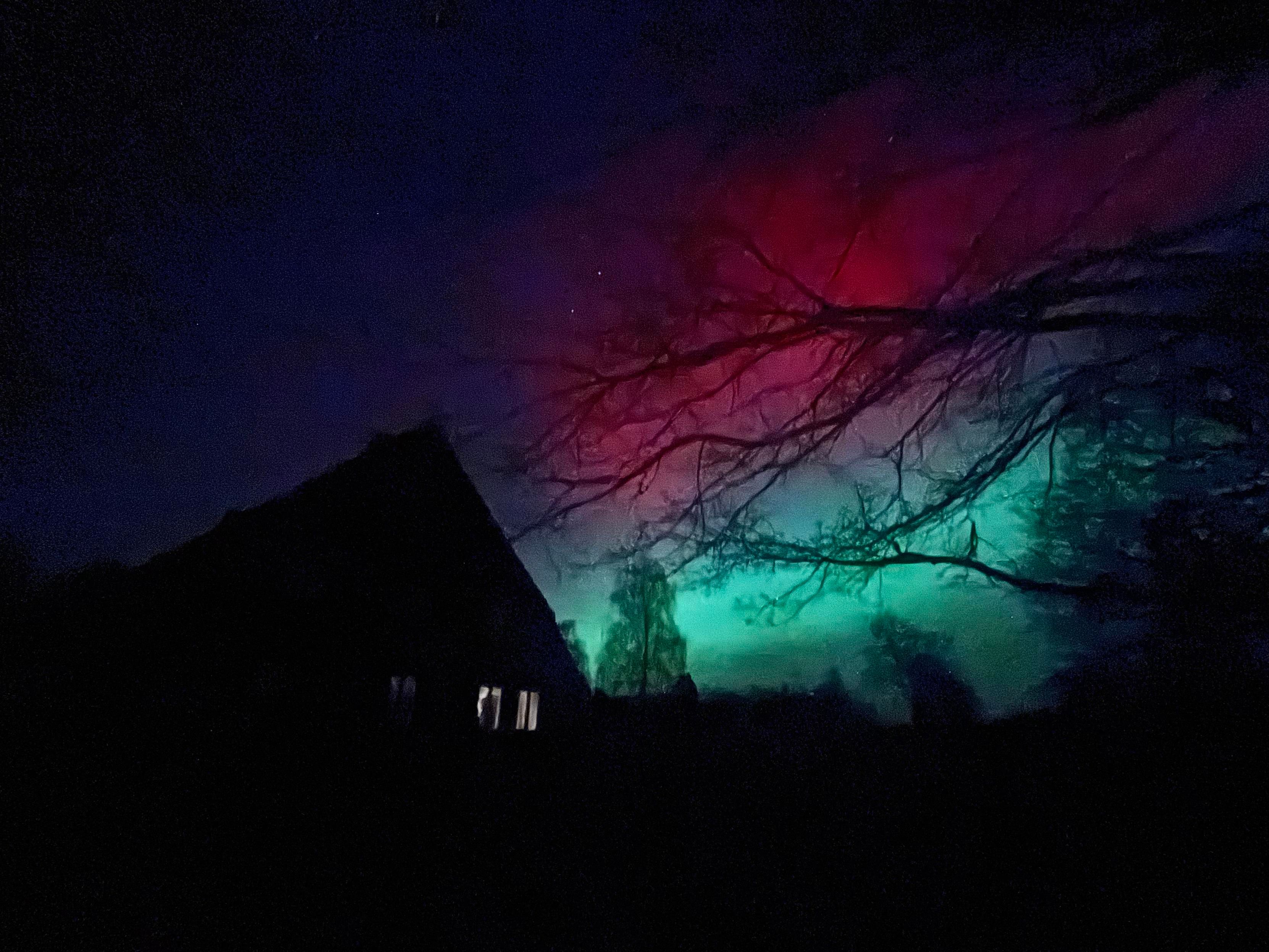 Eine dunkle Silhouette eines Bauernhauses vor einem bunten Nachthimmel mit leuchtenden Rot- und Grüntönen. Äste von nahe gelegenen Bäumen sind im Vordergrund sichtbar.