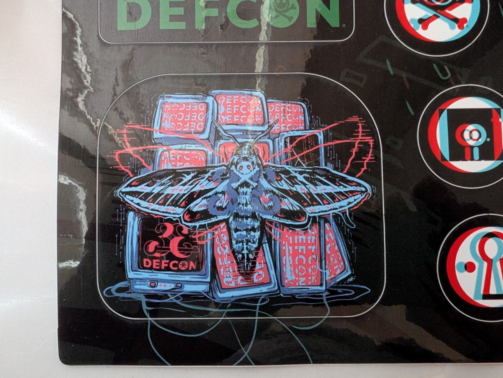 Illustration of a mouth fluttering around a cluster of monitors, all of which read "DEFCON.". On the moth's back is the DEFCON smiley-face skull and crossbones logo.