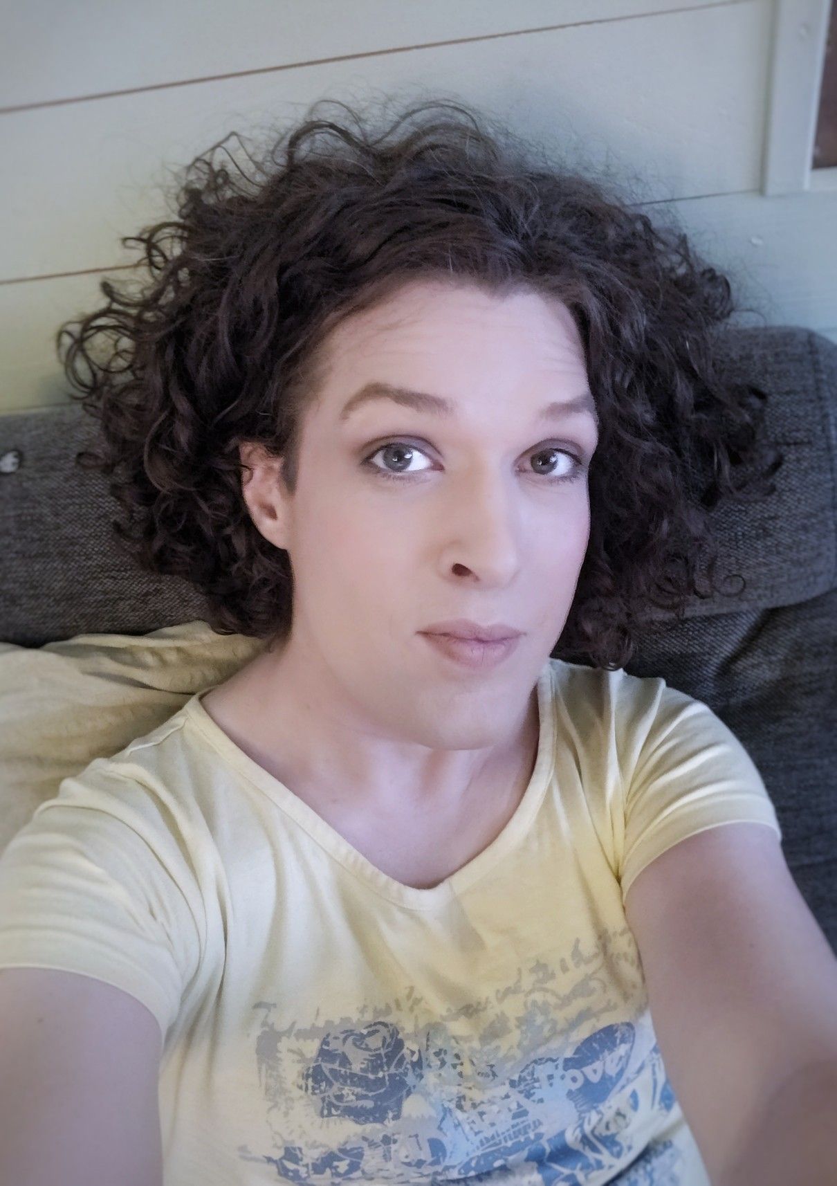 Selfie in sofa, wearing a yellow top, looking into the camera with a subtle smirk and lifted eyebrows, the hair tucked behind the ear on my right side.