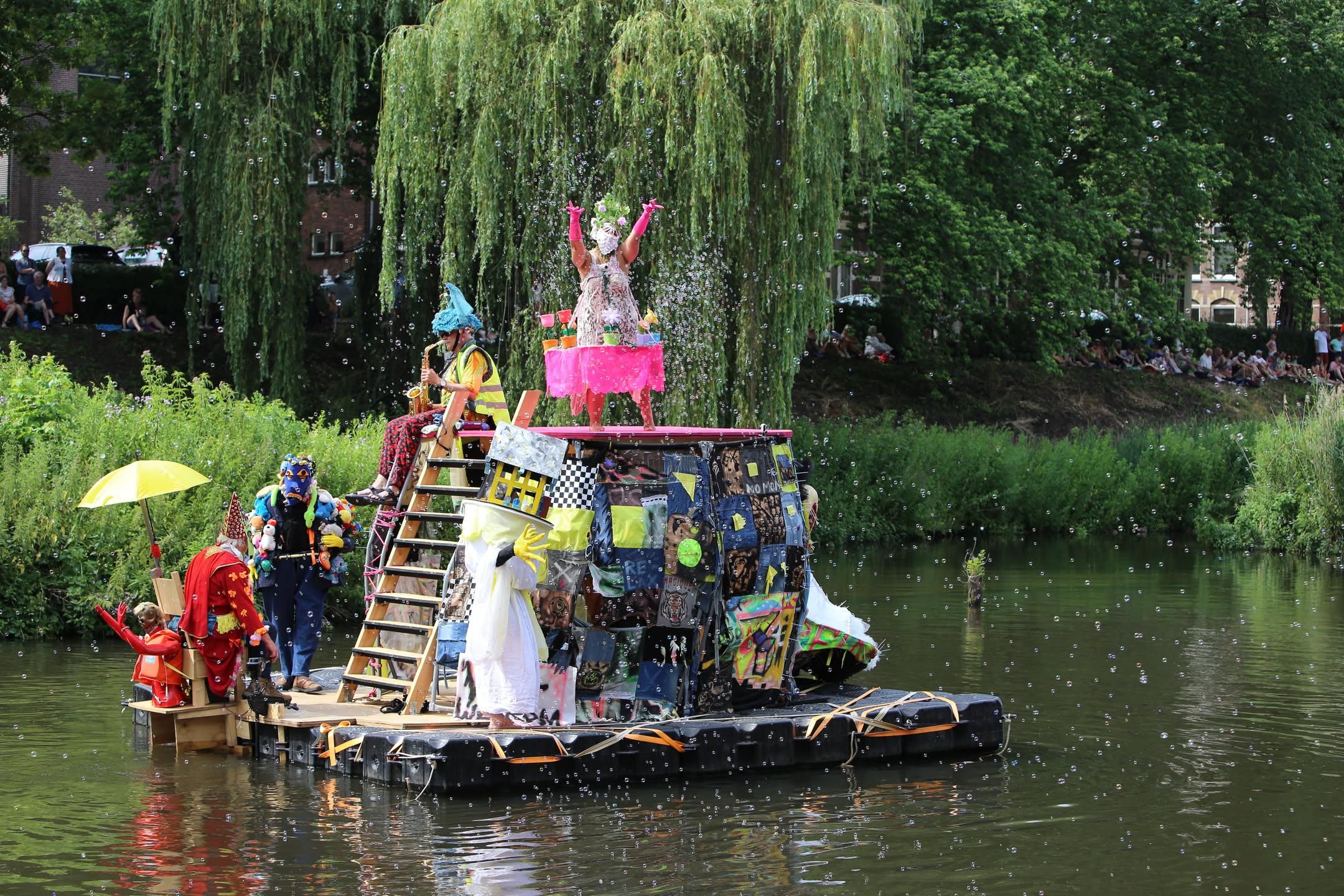 People in odd costumes dancing on a raft with a house that looks like a head. one is disgorging bubbles, another two are dressed like eyeballs for the head and a fourth is wearing odd yellow claws.