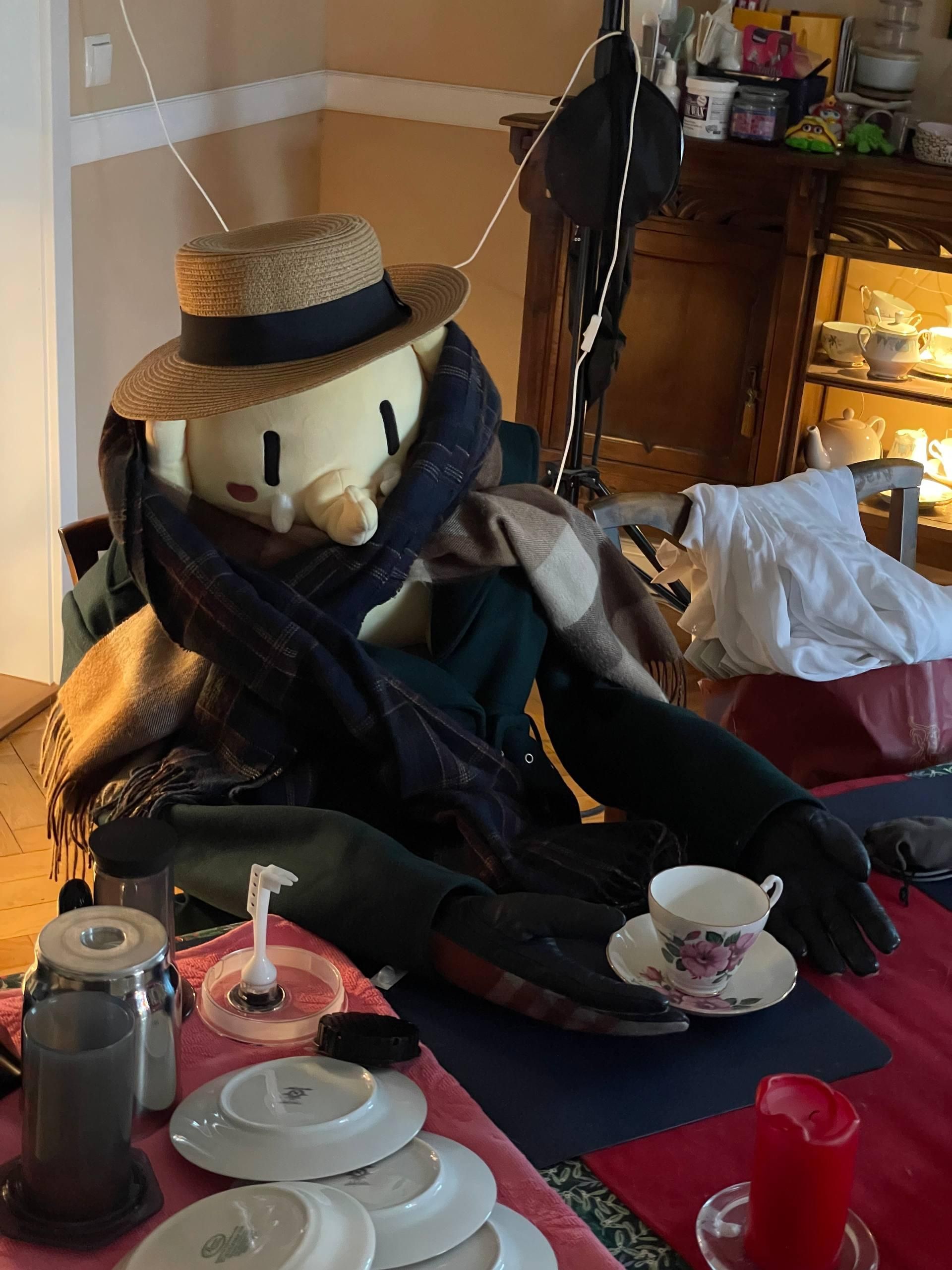The #Plushtodon XL sitting at the dinner table while wearing a coat, a scarf, a hat, and gloves, resembling a person, with an empty tea cup in front.