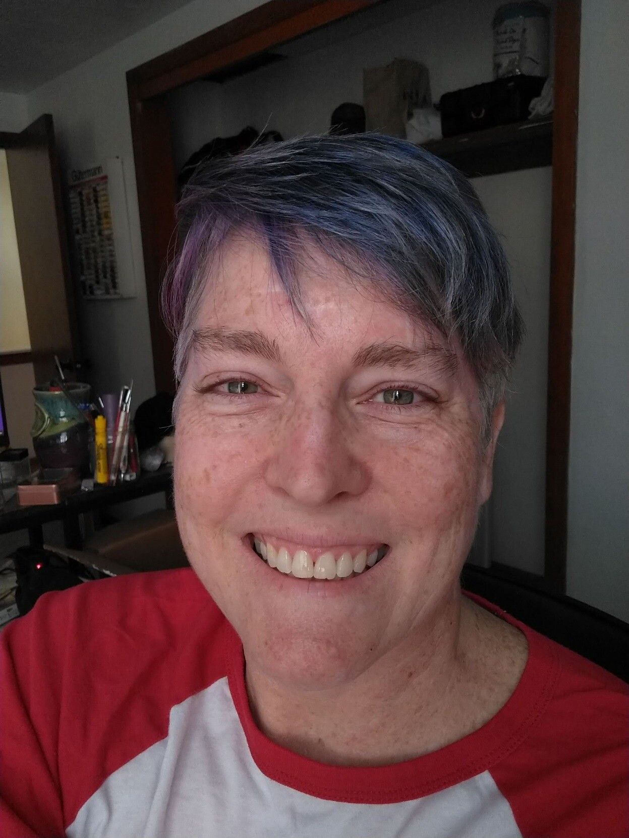 Selfie of a middle aged white lady with freckles, green eyes, and short blue and purple hair.