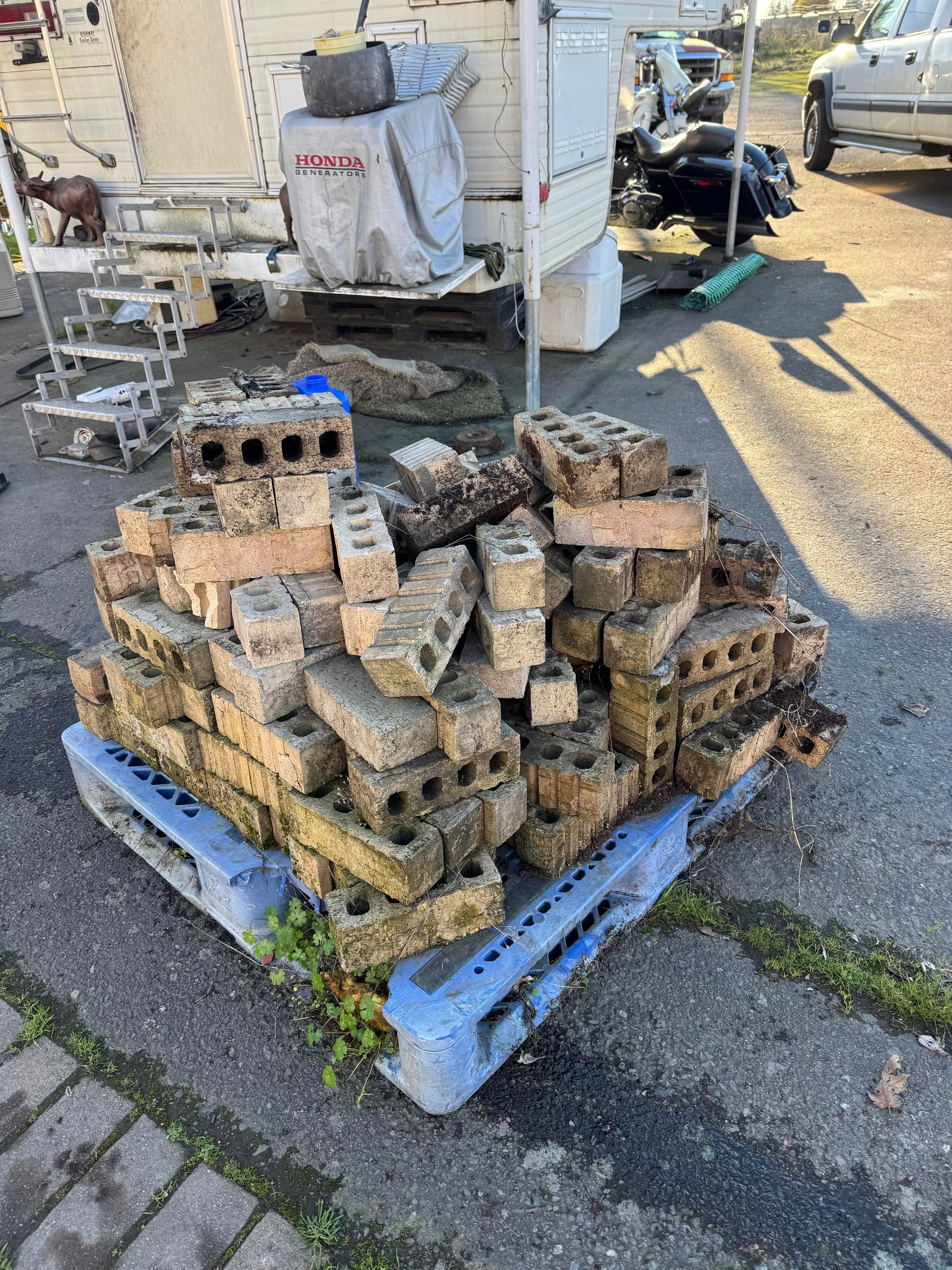 A stack of various concrete bricks and blocks is resting on a blue pallet, with a roadside setting that includes a travel trailer and parked vehicles in the background.