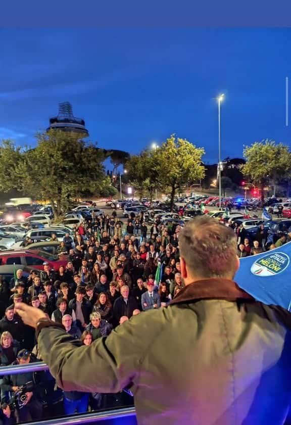 Foto del presidio dal palco su un bus. In un parcheggio, tra due file di auto parcheggiate, di vedono forse un centinaio di persone ad ascoltare.
