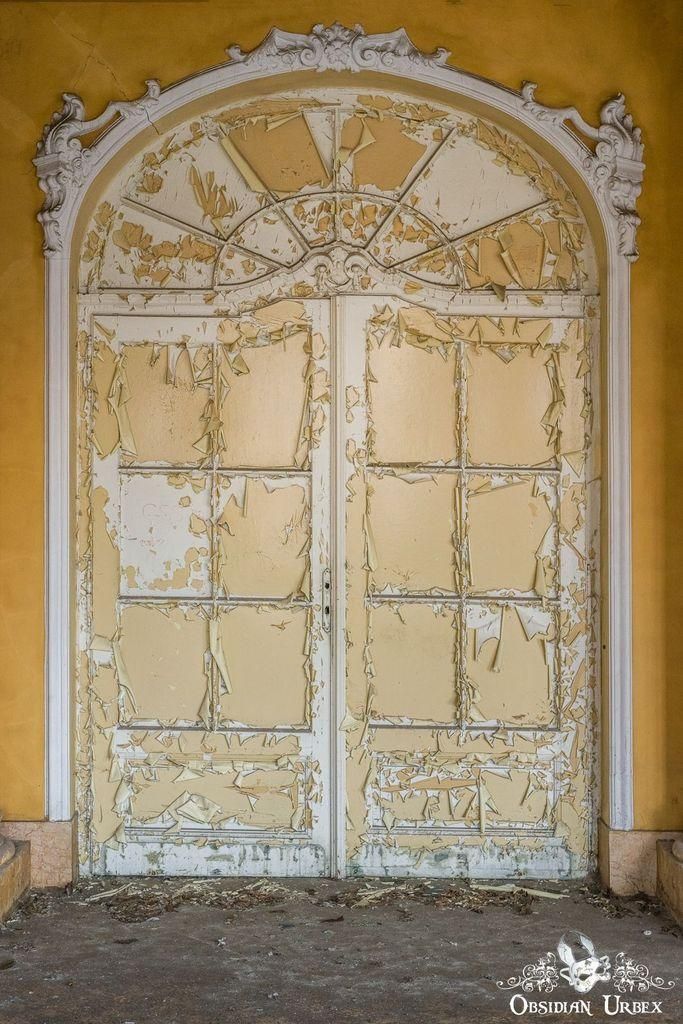 An ornate, arched doorway with peeling yellow paint, contrasting with a yellow wall. The door's decorative frame and decaying paint emphasize the theme of decay.