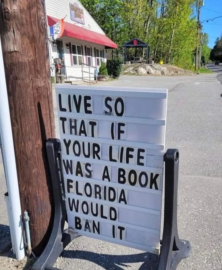 A roadside sign in front of a small building displays the message: 'Live so that if your life was a book, Florida would ban it.' This humorous and provocative statement refers to recent book bans in Florida, where certain books and topics have been restricted in schools and libraries due to their content. The sign encourages viewers to live boldly and embrace individuality, subtly critiquing censorship and advocating for freedom of expression. The setting includes a quaint building with a red awning and a quiet, tree-lined street in the background, adding a touch of small-town charm to the message.