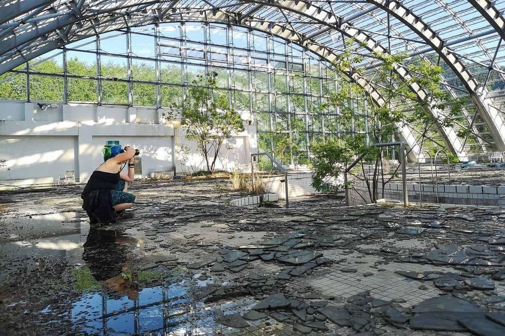 A person with a camera kneels on the cracked, debris-strewn floor of an abandoned glass greenhouse with overgrown plants and broken tiles.