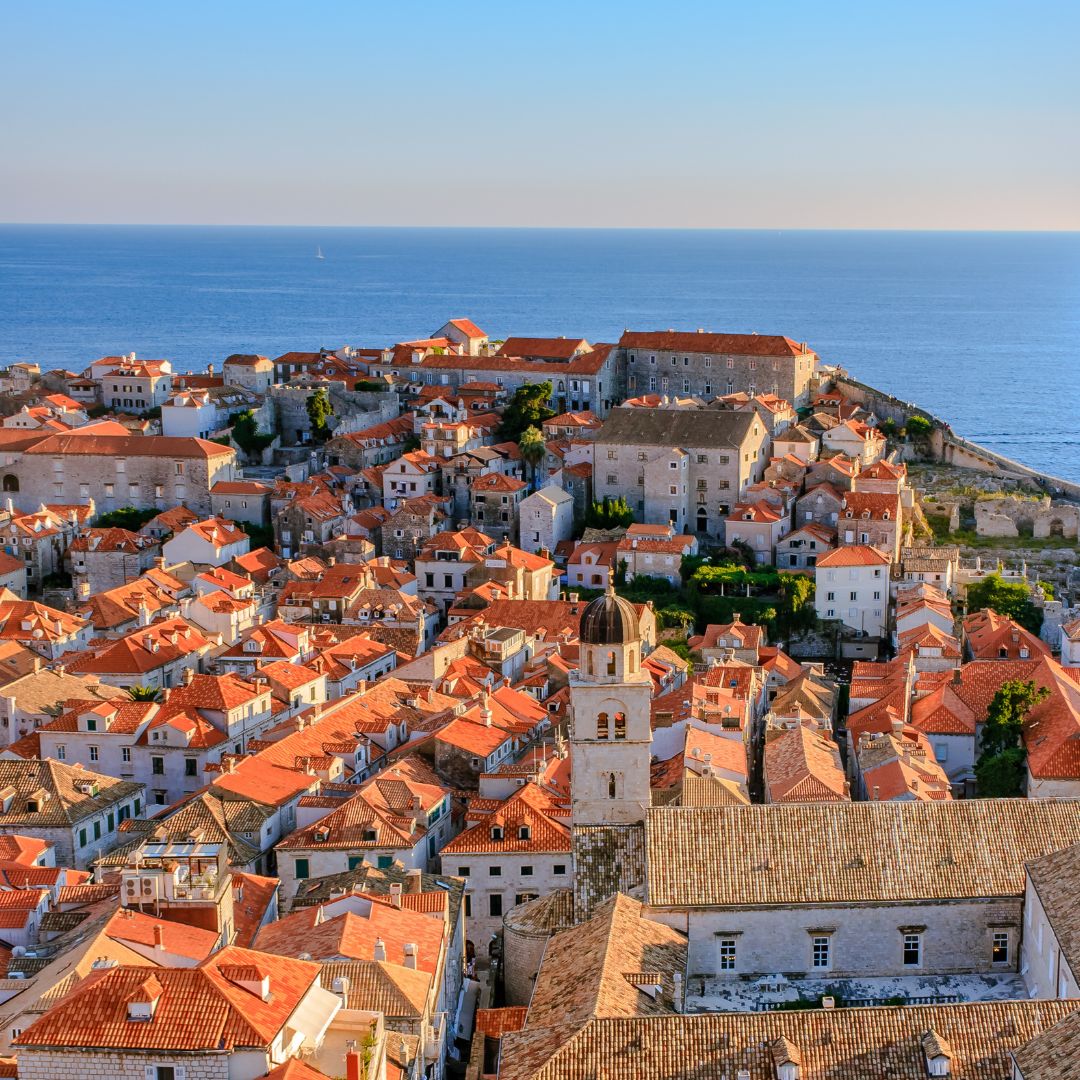 An aerial view of Dubrovnik with tightly packed stone buildings and terracotta rooftops. The town is nestled along a deep blue sea, with medieval walls and fortifications visible near the shoreline.