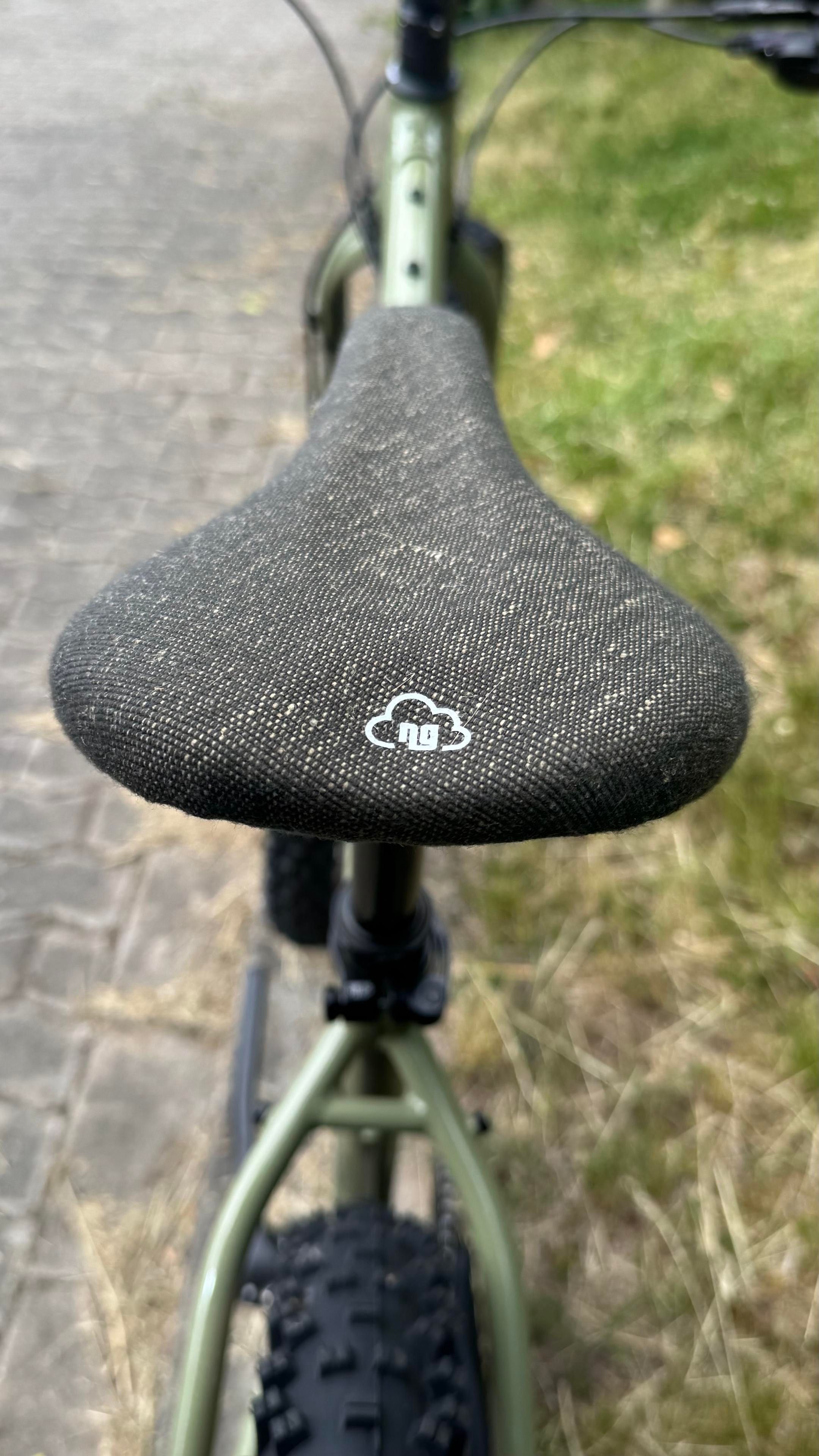Eine Nahaufnahme eines Fahrradsattels mit einem strukturierten schwarzen Bezug, mit einem weißen Logo, das einer Wolke ähnelt. Der Hintergrund zeigt Teile des Fahrradrahmens und des Bodens, was auf eine Außenumgebung hinweist.