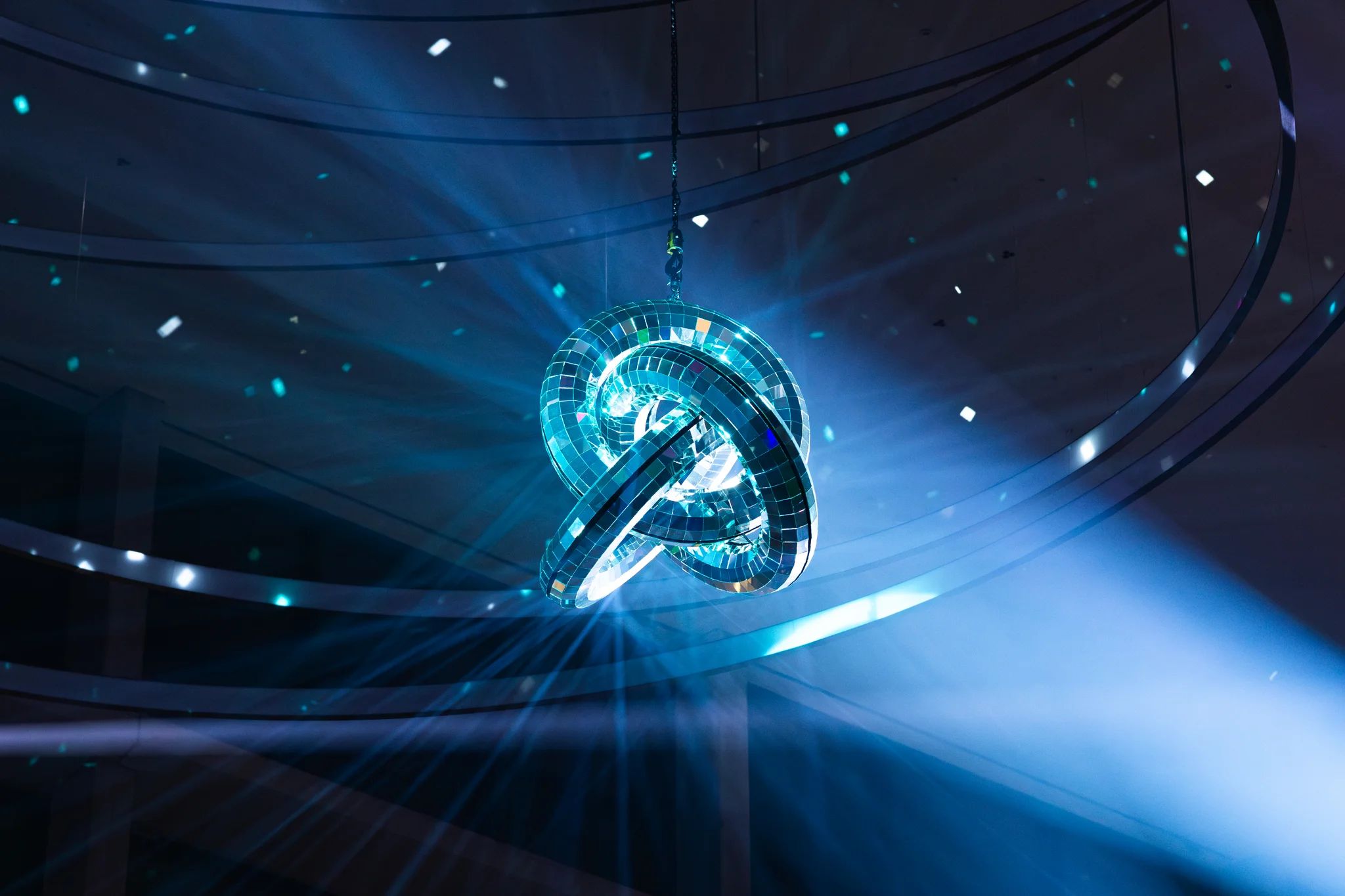 a disco ball-like structure consisting of 3 intertwined rings is refracting a light blue spotlight pointed onto it, causing little reflections to appear all over the big entrance hall's ceiling