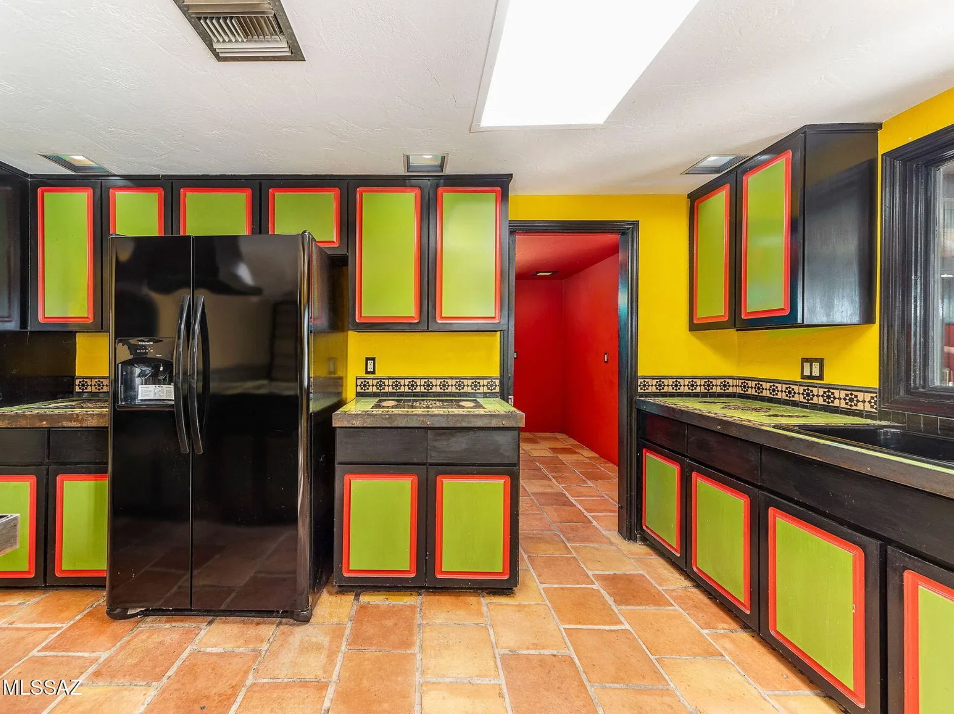 A kitchen with cabinets that are lime green with orange around the perimiter, and the sides of the cabinet are black, as is the fridge. The floor is pale orange tile. 