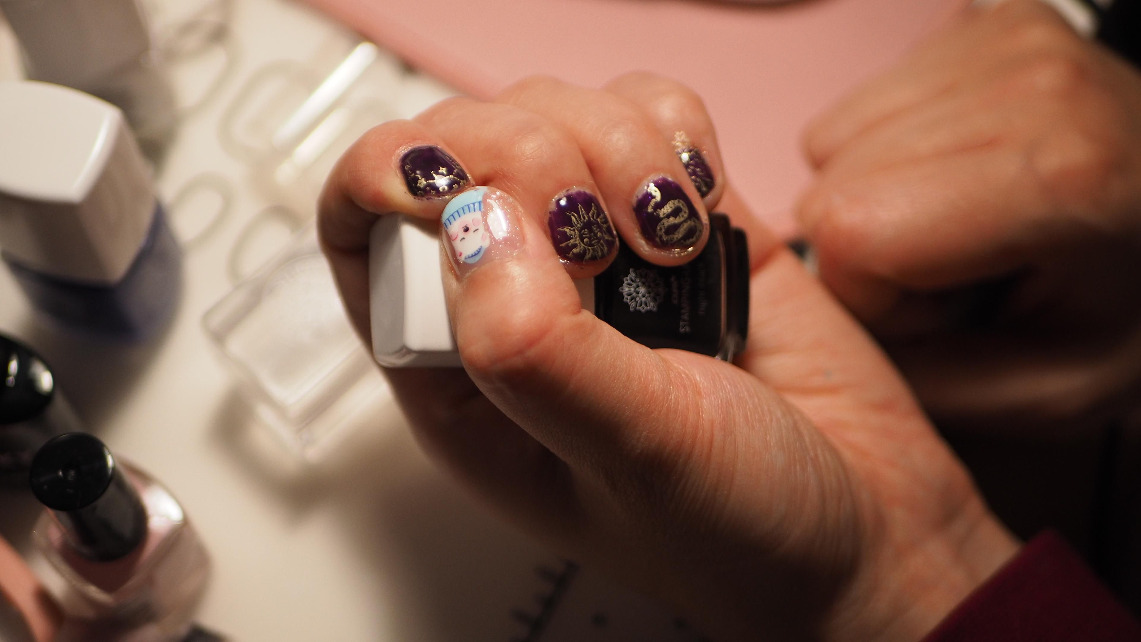 Another hand holding nail polish, the thumb nail on this one displays a cartoon bear while the other fingers have a dark background with golden motifs of stars, the sun, and a snake.