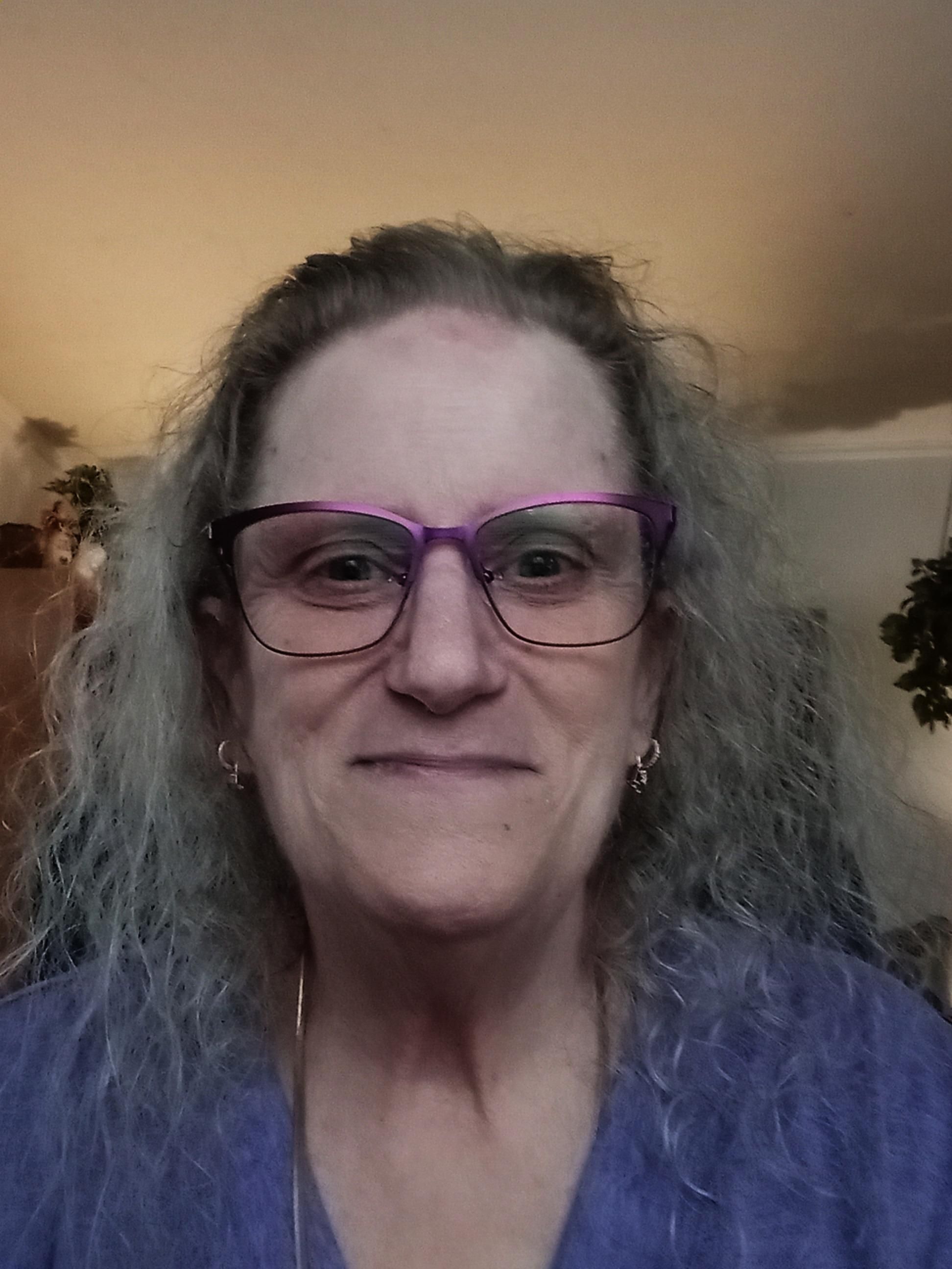 Selfie of a woman (me). I'm wearing purple eye-glasses and a purple shirt. I have long-ish curly hair - it used to be blonde, now it's blond adjacent.