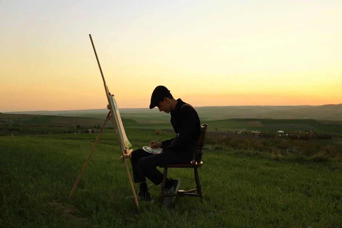 A masc figure sits on a wooden chair in a field, in front of an easel, with a paint palette in their lap. They're wearing black with a black peaked cap. The sky behind them is pastel orange.
