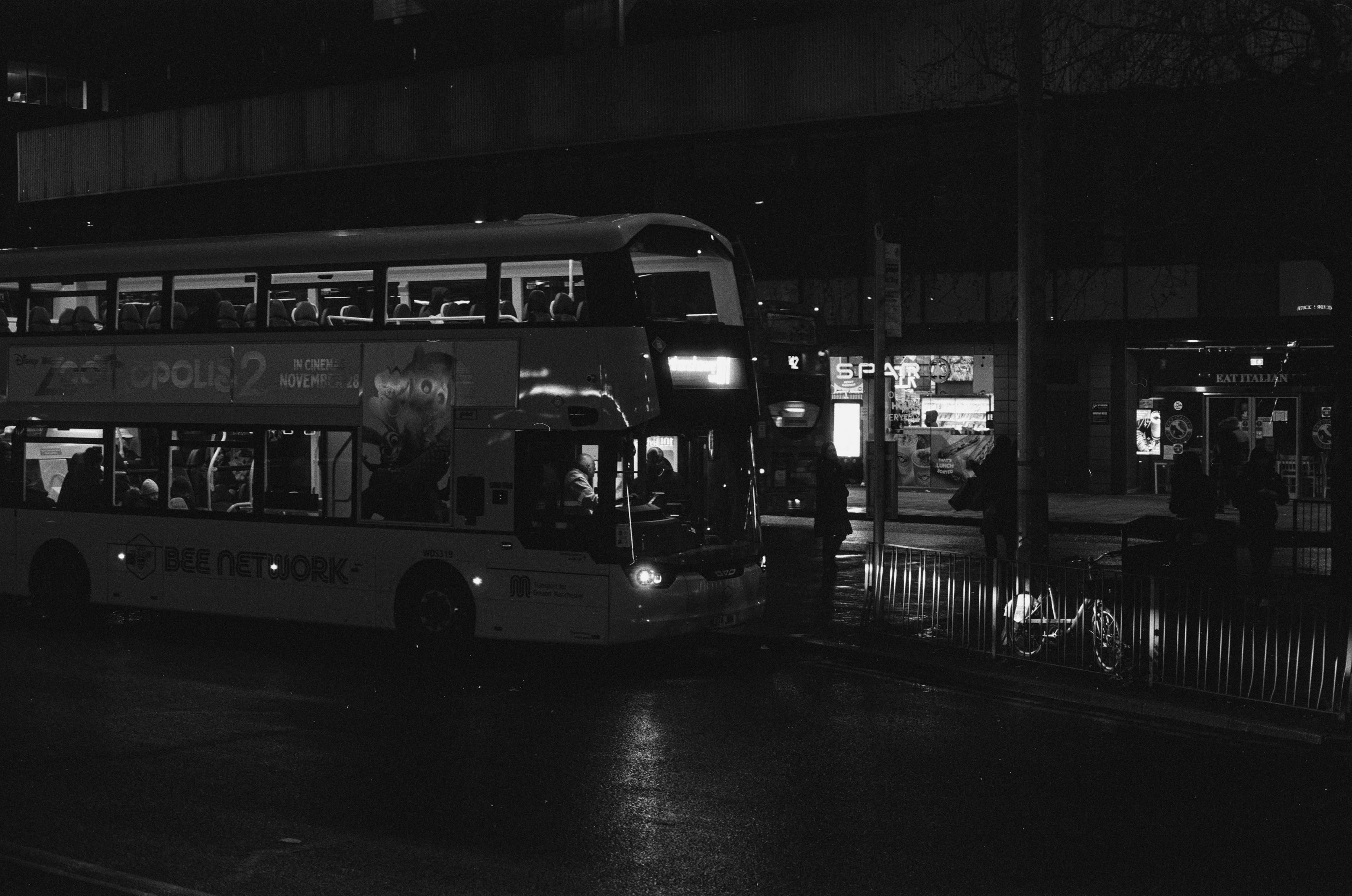 A double decker Bee Network bus parked at Piccadilly Gardens with a SPAR visible behind it at night. The asphalt is wet from rain and the bus lights are reflecting off of a metal fence in front of it.