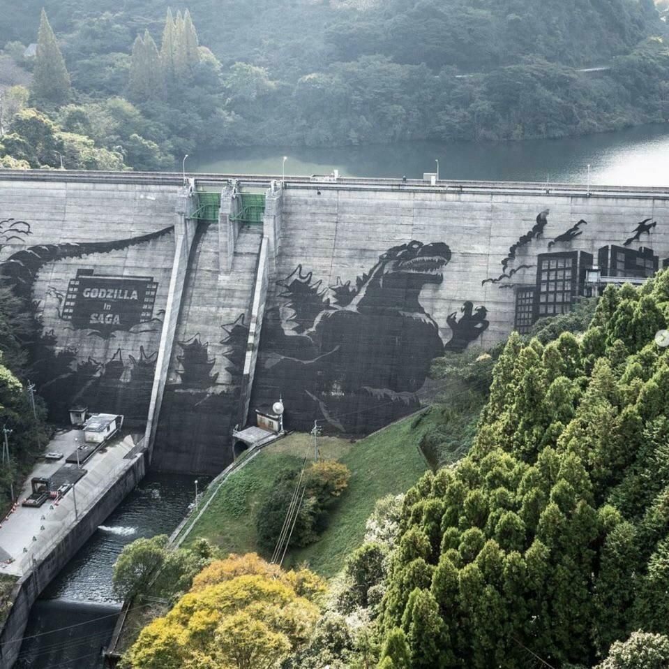 A photograph of a huge dam, an aerial view. The dam has been power cleaned leaving a relief of Godzilla, with the words Godzilla and Saga above. To one side and behind green hillsides and trees flank the lake that the dam holds back.