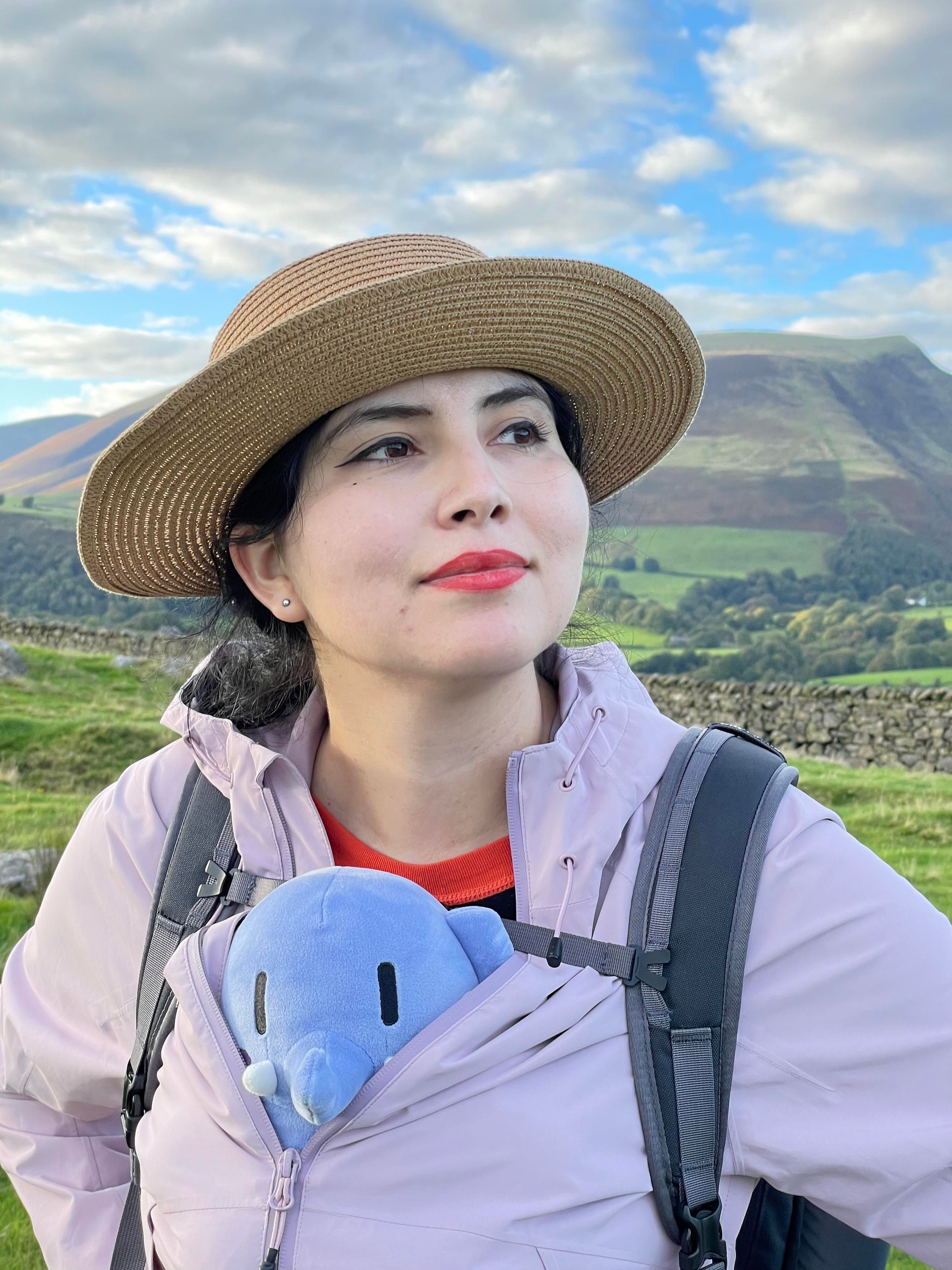 My wife dressed in outdoor clothing with a blue stuffed Mastodon toy peaking out from her jacket in front of a lusciously green landscape. 