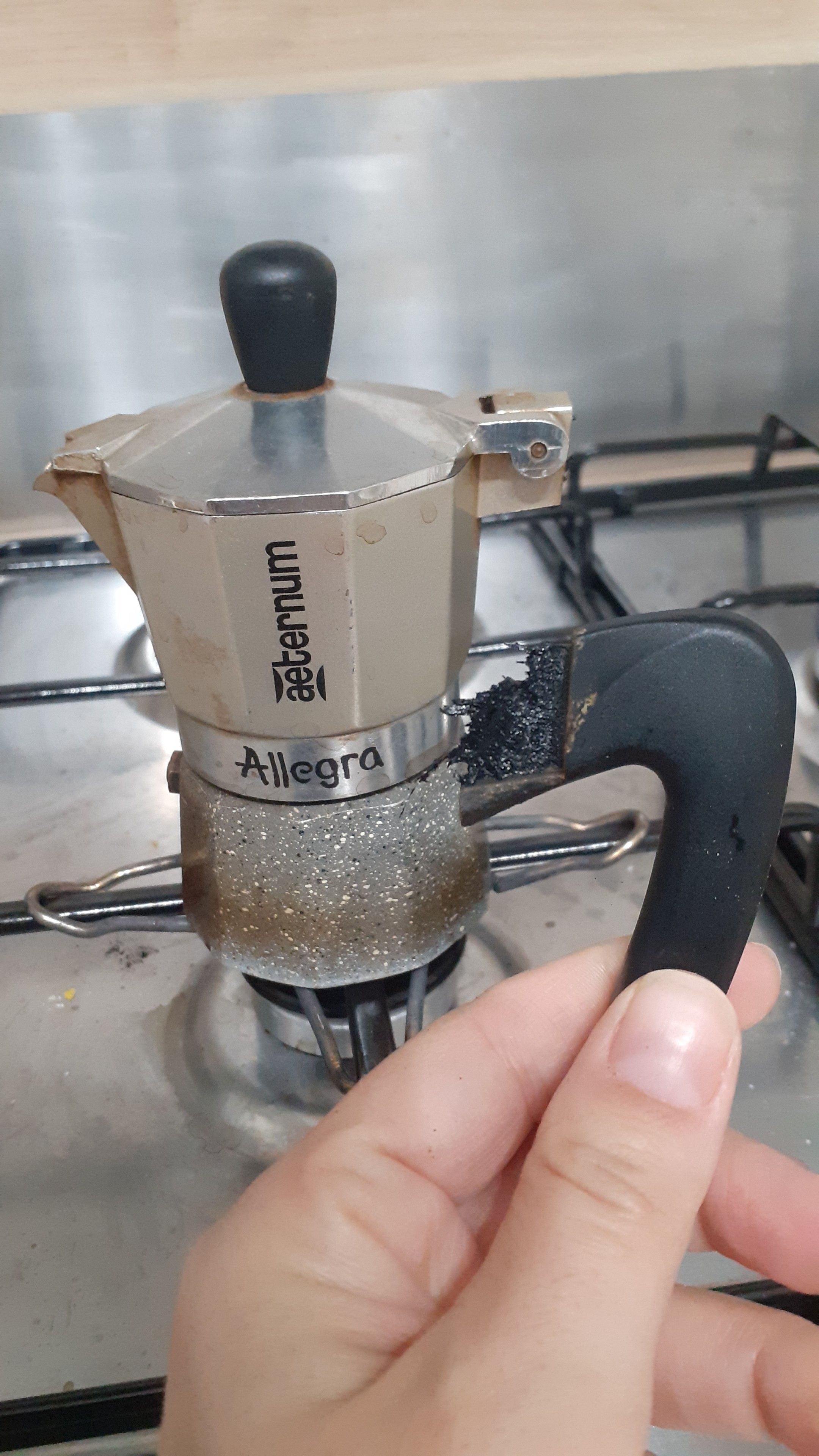 la mia mano che tiene il manico della moka piccola che si è staccato perché si è sciolto nel punto in cui si congiungeva alla moka. sullo sfondo la moka, sopravvissuta, che adesso sa di bruciato
