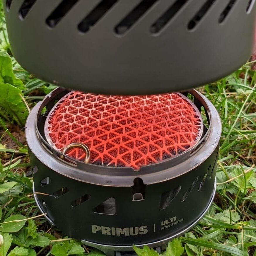 Stock image of a Primus brand catalytic camp stove. The stove sits on green vegetation and a heat exchanger pot hovers above the stove held by an unseen hand. The stove glows bright orange.