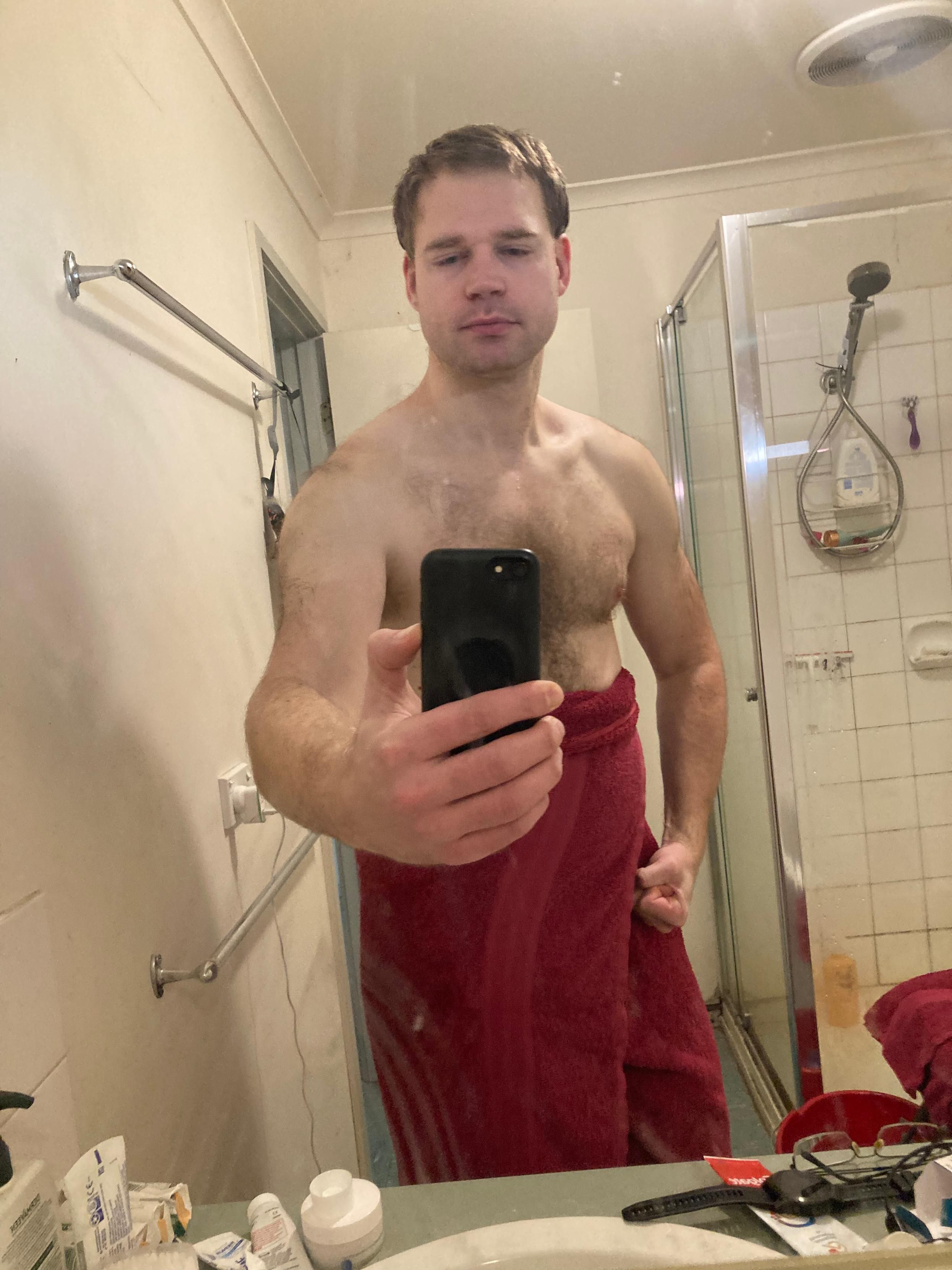 post-shower bathroom selfie of Luke. They are smiling at the camera and showing off their chest. They are wearing a red bath towel around their waist.