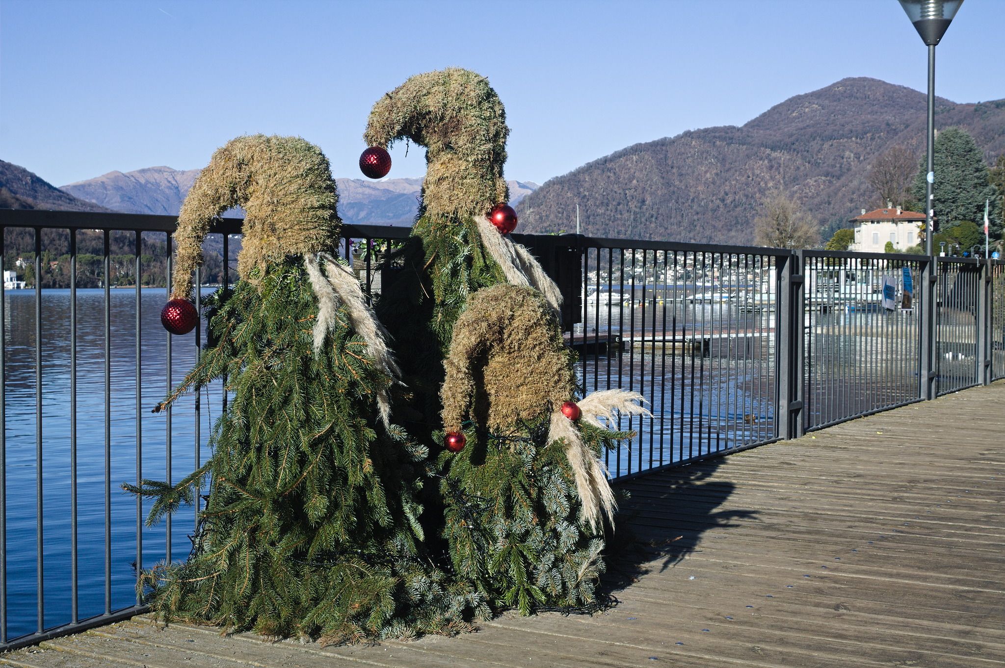 three gnome adjacent creatures made of branches of evergreen trees, with a pointy hat made of moss, big white moustaches and red Christmas tree balls at the end of the hat and as a nose. They are wrapped in LED strings, and the lake can be seen in the background.